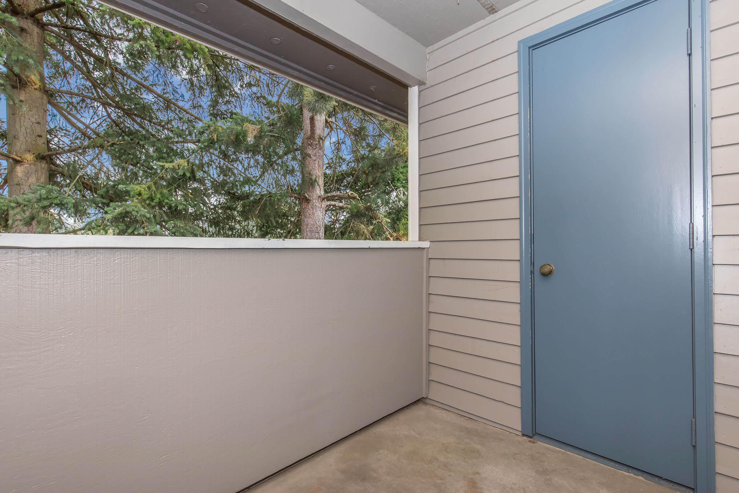 A small outdoor balcony or porch area featuring a light-colored wall, a blue door, and visible trees outside through a screened window. The concrete floor adds a modern touch, creating a cozy and inviting space for relaxation.