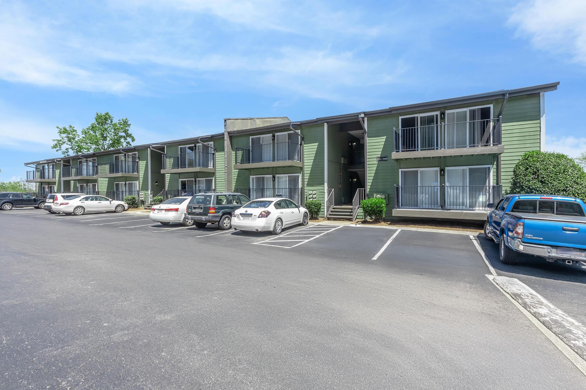 A two-story green apartment building with multiple entrances, surrounded by parked cars in a paved lot. The sky is clear and blue, with a few trees in the background. The building features balconies and stairs leading to the upper level.