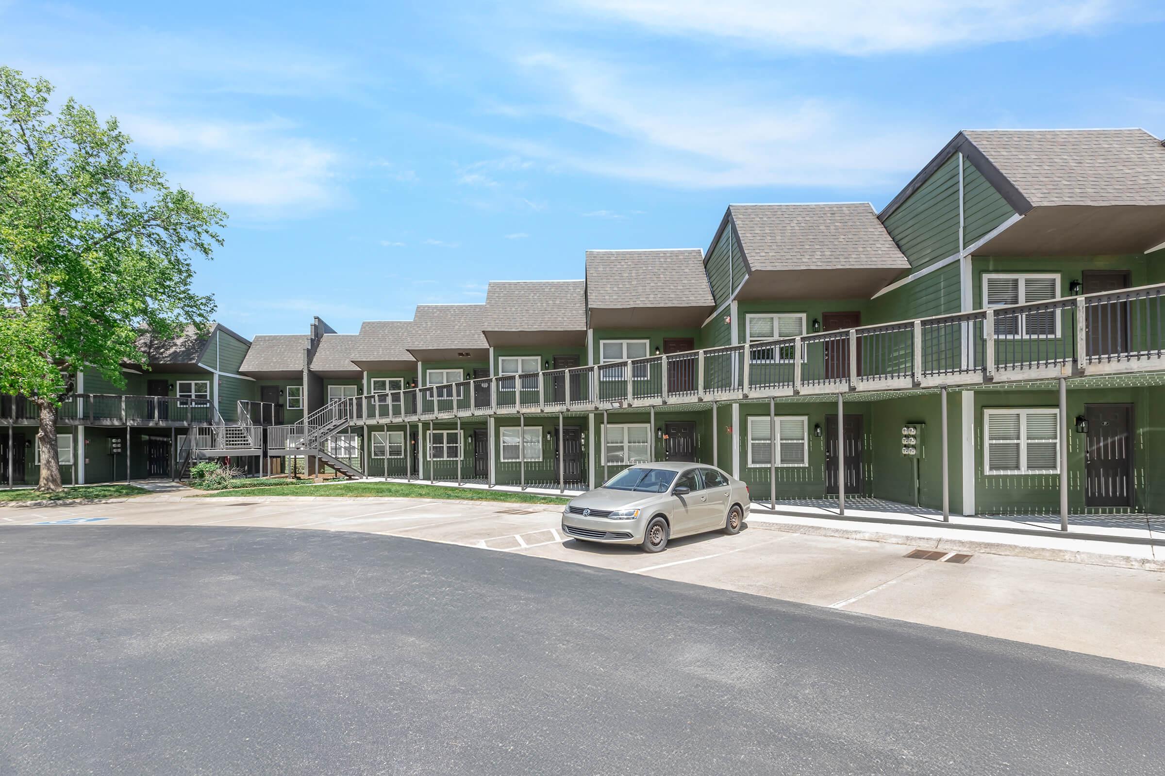 A photo of a green apartment complex featuring multiple two-story buildings with balconies. The parking lot is visible in front, with one silver car parked. Trees are present in the area, and the sky is clear. The overall scene conveys a serene residential environment.