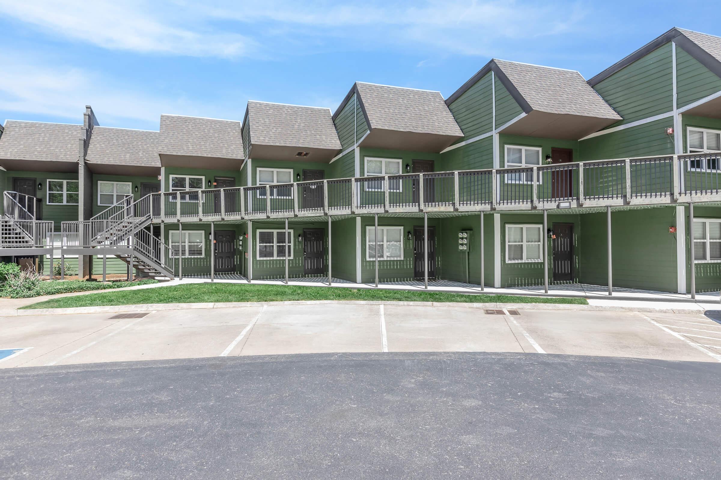 Row of green apartment buildings with multiple units, featuring balconies and staircases. The exterior has a modern design with triangular roofs and large windows. A parking area is visible in front, with clear skies above and landscaped grass surrounding the buildings.