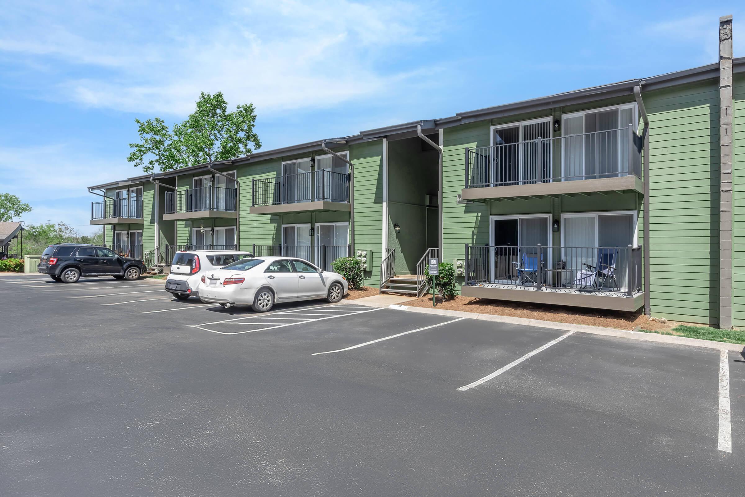 Two-story green apartment building with balconies, surrounded by a parking lot. Several cars are parked in front, and there are trees in the background under a clear blue sky. The scene is inviting and well-maintained, suggesting a residential area.