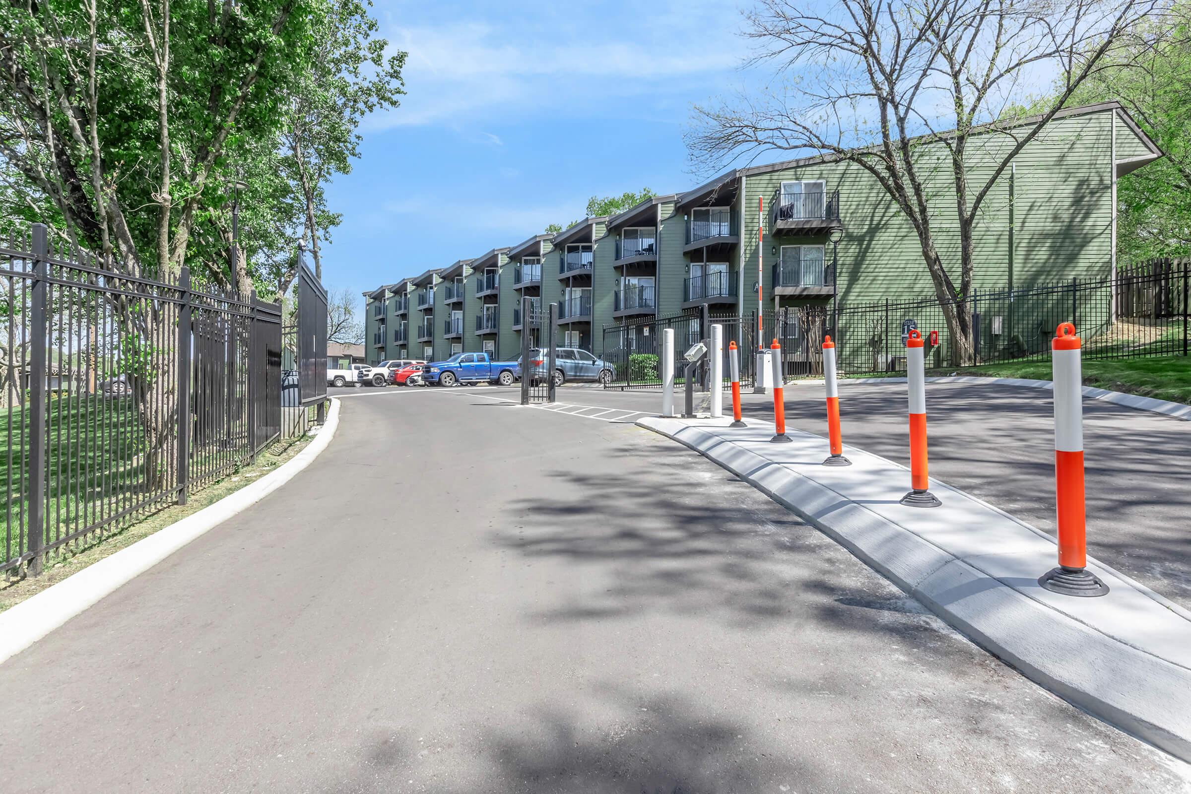 A view of a paved access road leading to an apartment complex with multiple units. There are orange and white traffic cones marking the road, which is bordered by trees and a black fence. Several parked cars are visible near the building, under a clear blue sky.