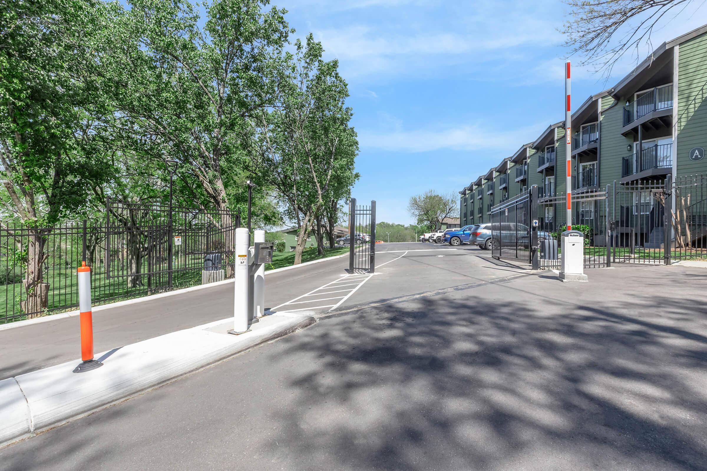 A gated entrance to an apartment complex, featuring a security gate with an access control system. The scene includes lush green trees on either side, a clear blue sky, and parked cars visible in the background. The pathway is well-maintained, leading to the buildings in the complex.