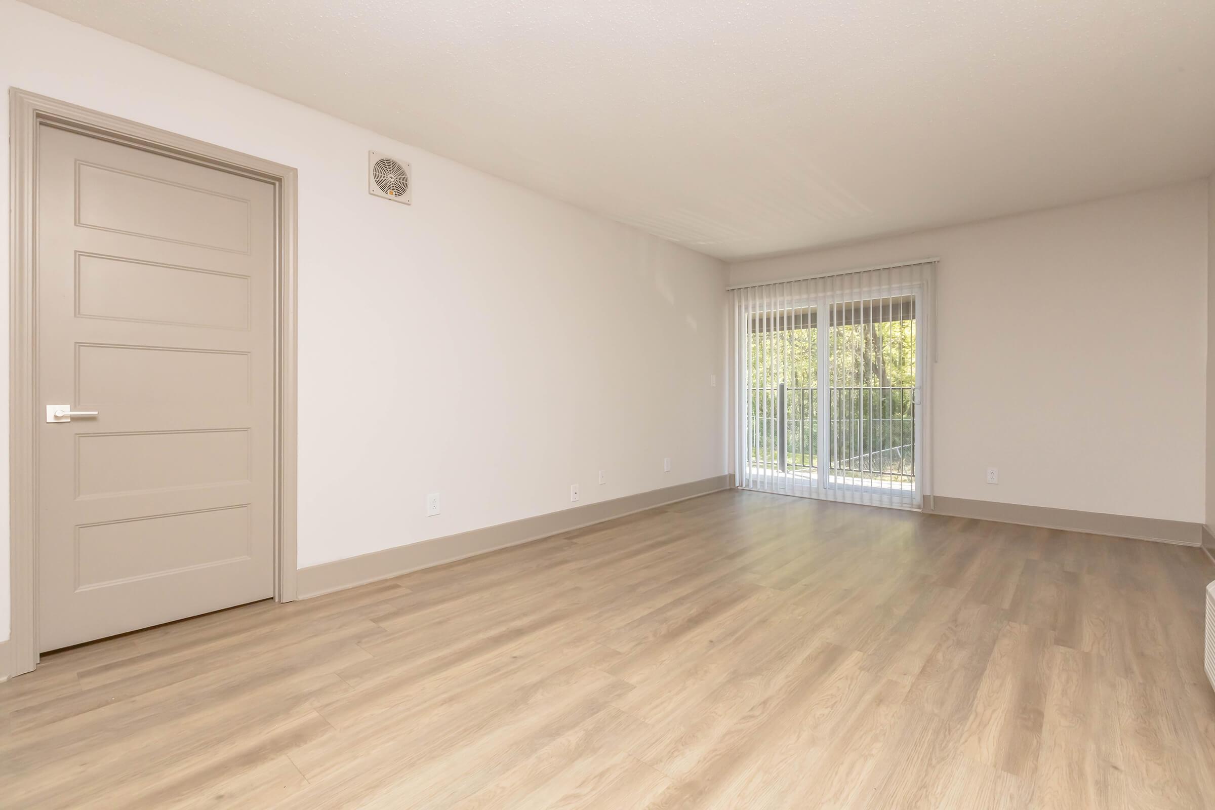 A spacious, empty room with light-colored walls and a wooden floor. A closed door leads to another area, and large windows with blinds provide natural light. The room features a minimalist design, emphasizing openness and potential for furnishings.