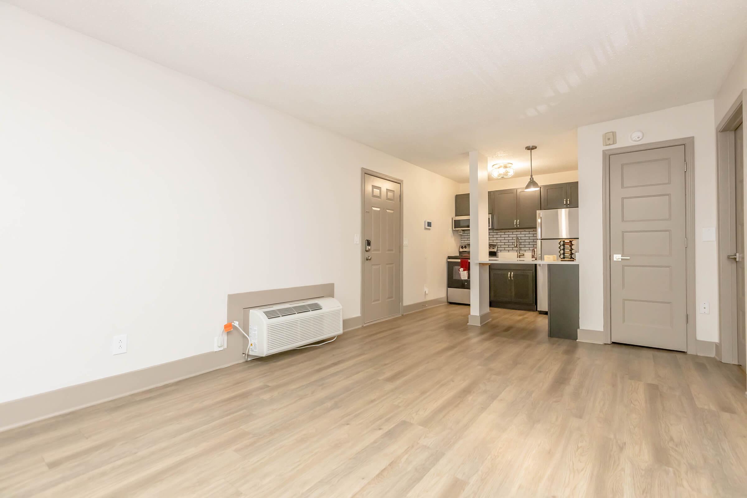Modern, empty apartment interior featuring a light-colored wall, wooden floor, and a small kitchen area visible in the background. There's a wall-mounted air conditioning unit and a door leading to another room. Natural light brightens the space, emphasizing its clean and spacious layout.