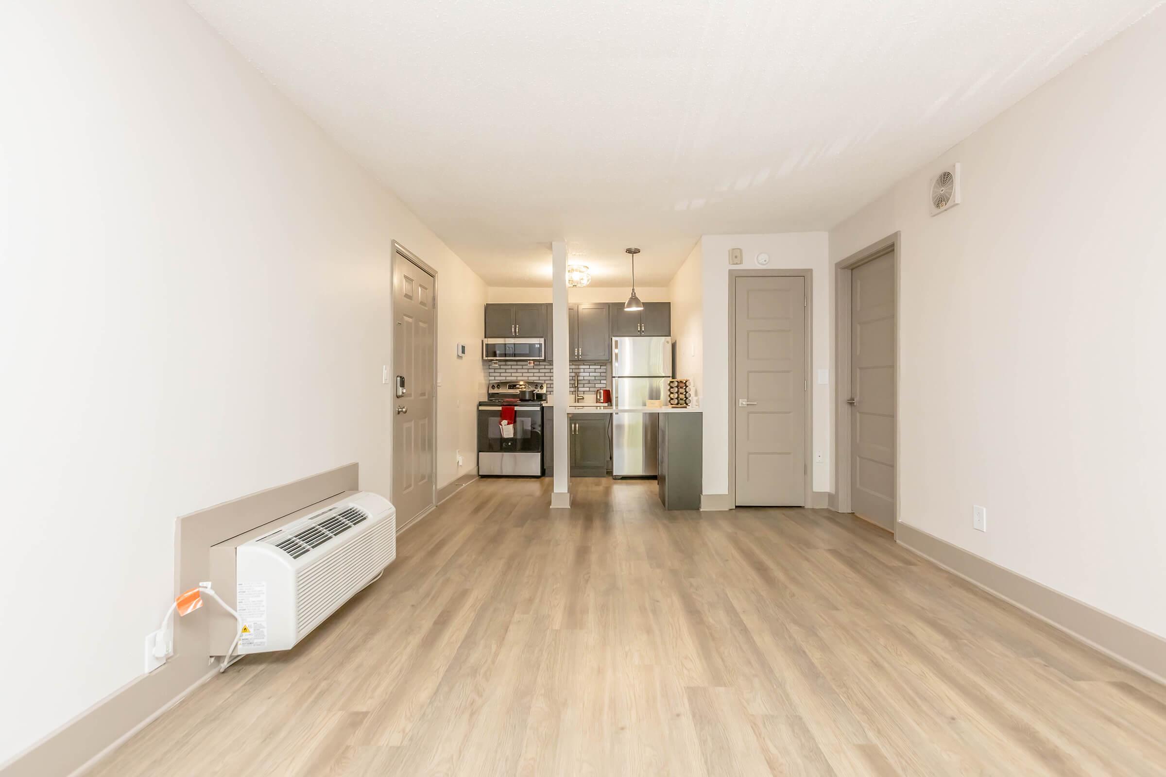 An interior view of a modern, empty apartment featuring a spacious living area with hardwood floors, a wall-mounted air conditioner, and a kitchen in the background. The kitchen includes stainless steel appliances and dark cabinetry, with a light fixture above the dining area. Soft lighting creates a welcoming atmosphere.