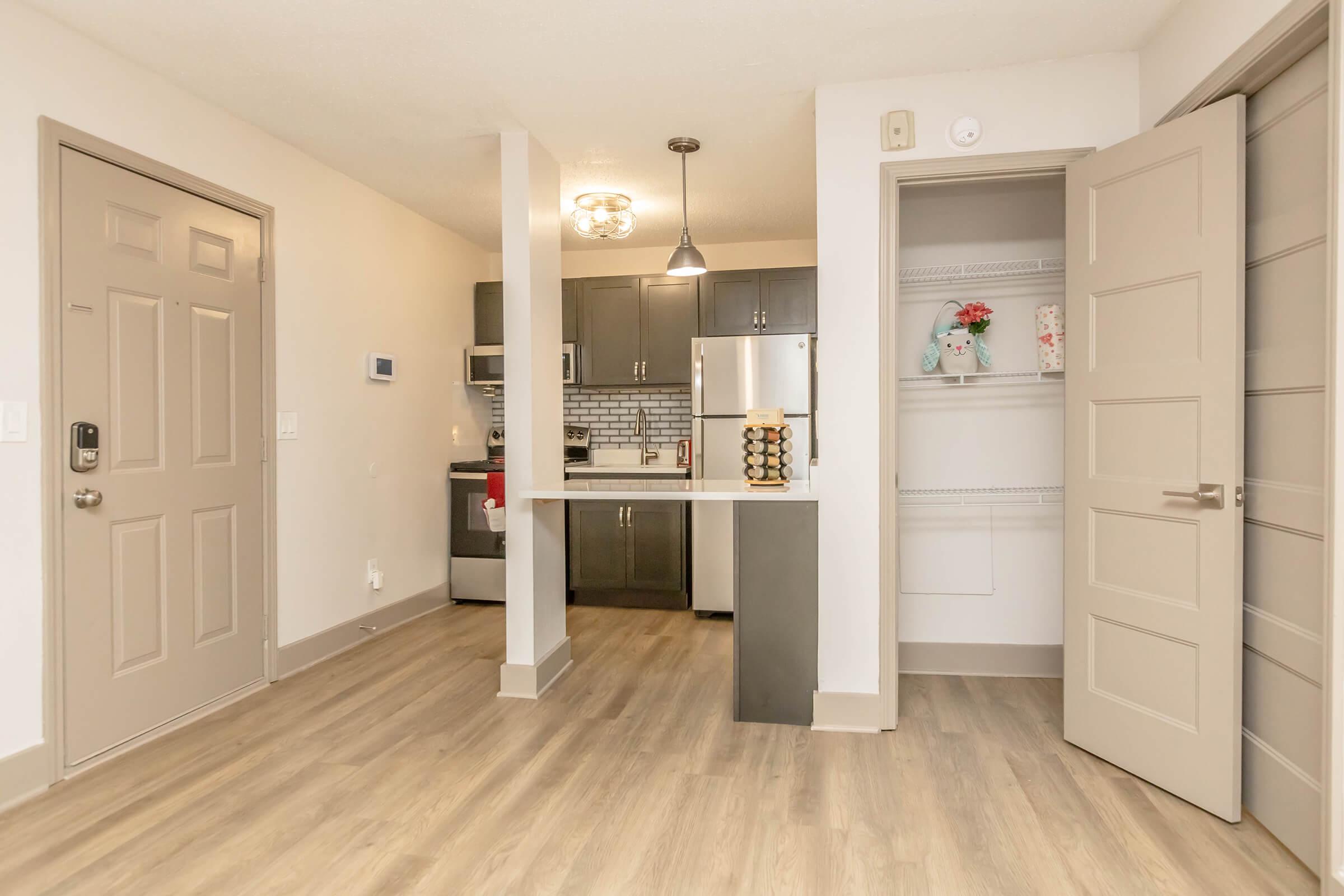 A modern kitchen and living area featuring an open layout. The kitchen has dark cabinetry, a silver refrigerator, and a window. There's a dining area with bar seating. A door leads outside, and a closet is visible on the right side. The space is well-lit with neutral-colored walls and wood-like flooring.