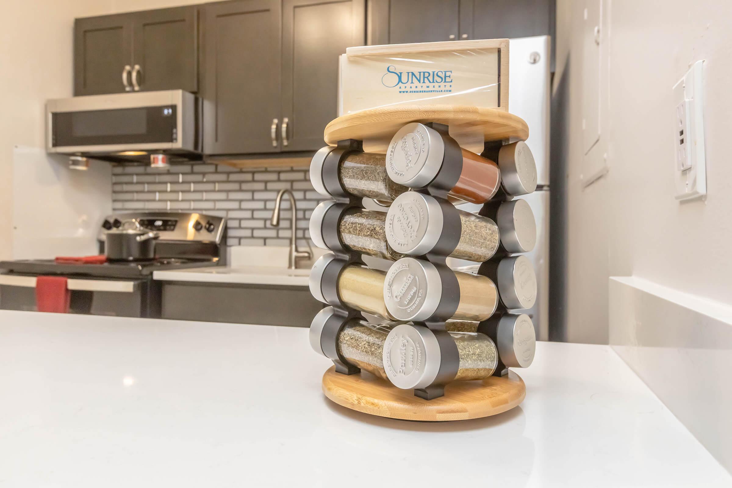 A rotating spice rack made of bamboo, holding multiple clear containers of various spices and herbs, displayed on a kitchen countertop. The background features dark cabinetry and a stainless steel microwave.