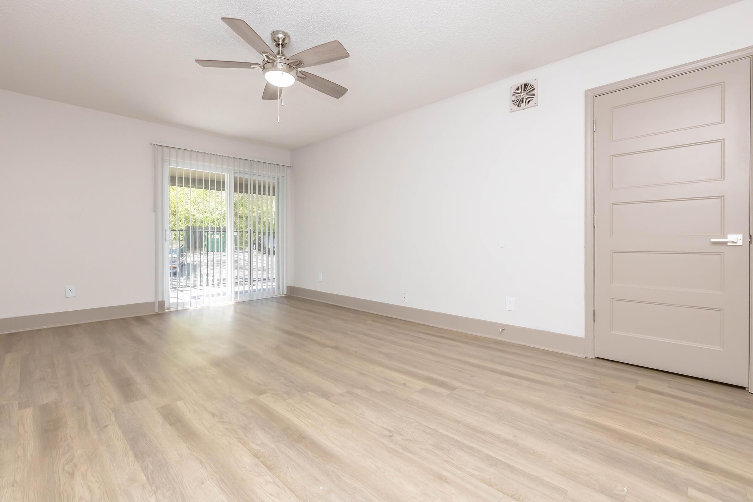 A spacious, light-filled room with a ceiling fan, neutral-colored walls, and smooth wooden flooring. It features a door leading to an outdoor space with vertical blinds, creating a bright and airy atmosphere. The clean and simple design allows for versatile furniture arrangement.