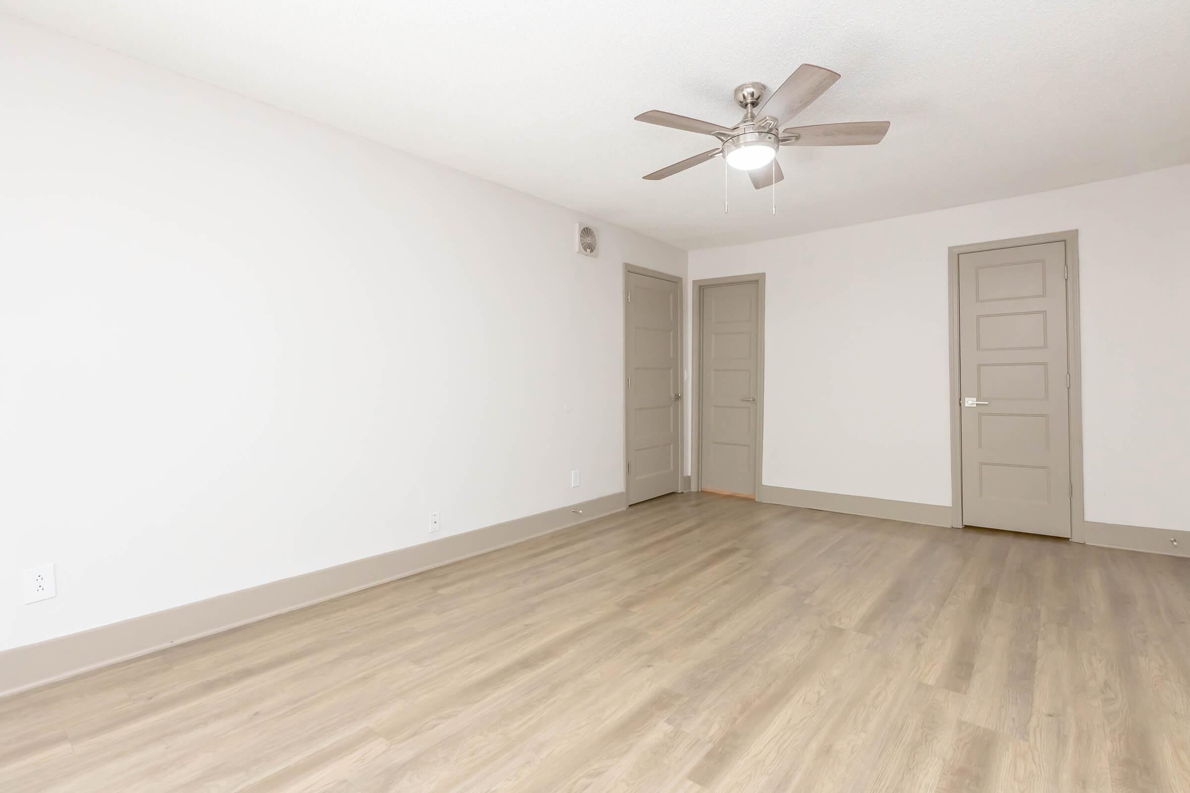 A spacious, empty room with light-colored walls and a ceiling fan. The floor is covered with light wood-like laminate. There are two closed doors on the right side, leading to other areas, and the room is well-lit with natural light. The atmosphere is clean and modern, ideal for various uses.