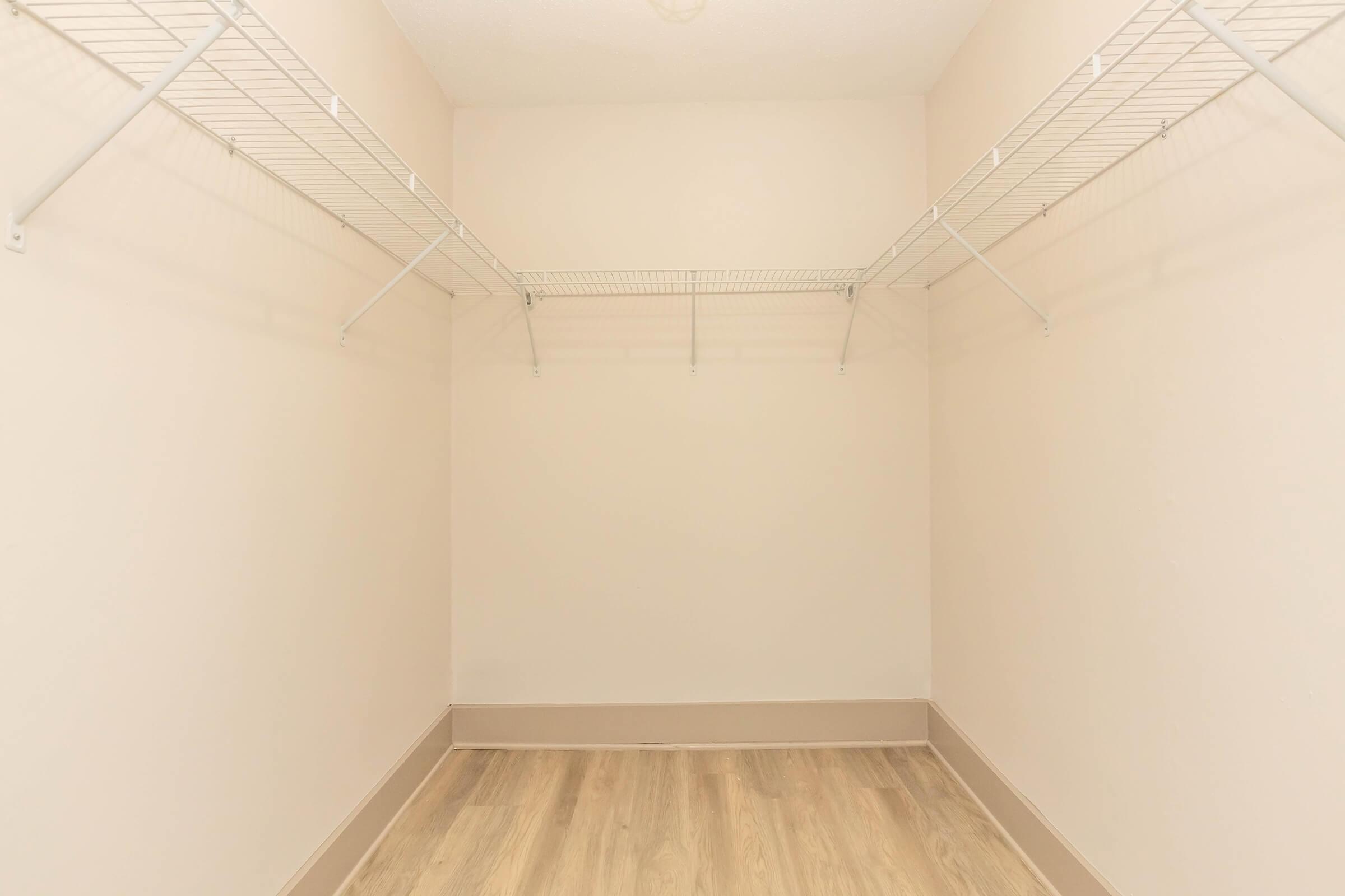 Empty closet with light-colored walls, featuring two wire shelves on either side. The floor is a wood-like material, and the overall space is well-lit and minimalistic.