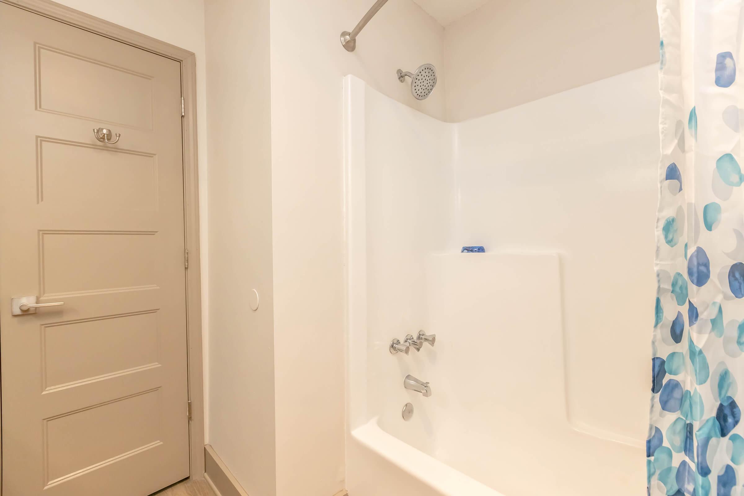 A clean bathroom with a white bathtub, showerhead, and a colorful shower curtain featuring blue and green polka dots. A light-colored door is visible on the left, providing access to the bathroom. The overall design is modern and minimalist, with neutral tones and bright lighting.