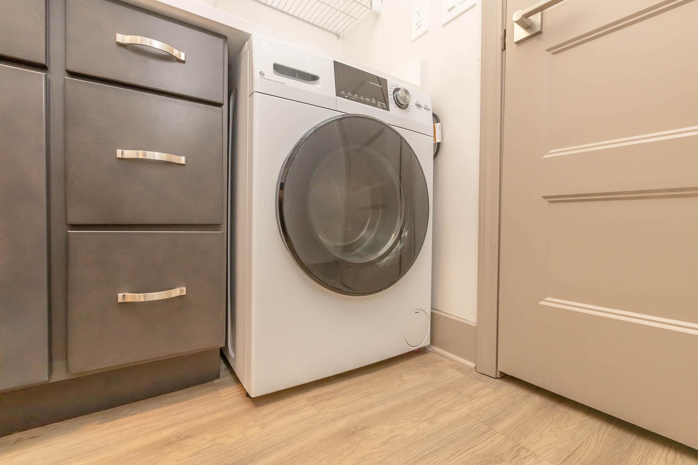 A modern white front-loading washing machine placed in a laundry room next to a dark cabinet. The floor is light-colored wood, and there's a door slightly ajar in the background. The setting appears clean and organized.