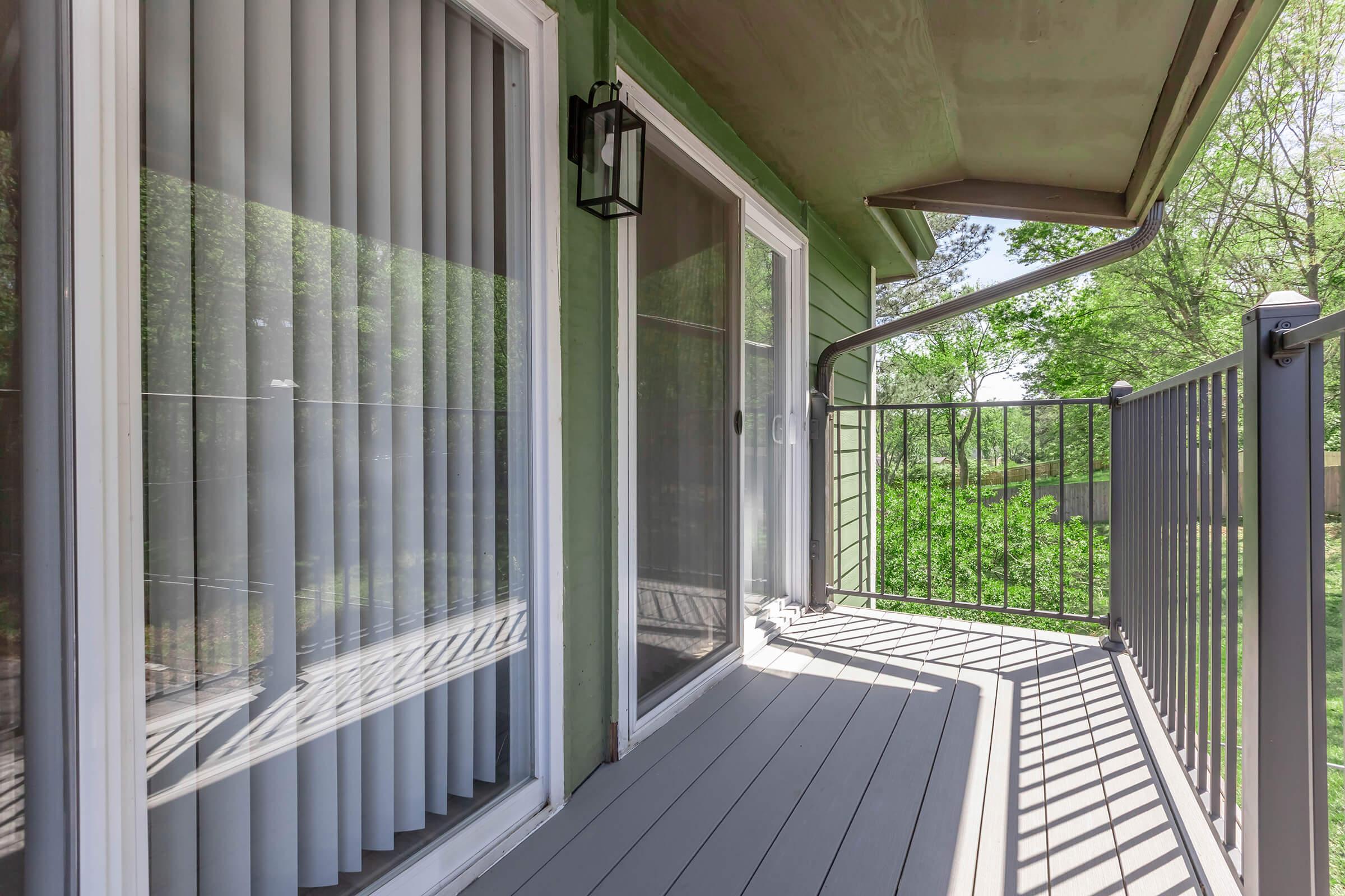 A modern balcony with a green exterior wall, featuring large sliding glass doors and vertical blinds. The balcony floor is made of gray decking, and a black railing encloses the space. Sunlight casts shadows on the floor, and greenery is visible in the background.