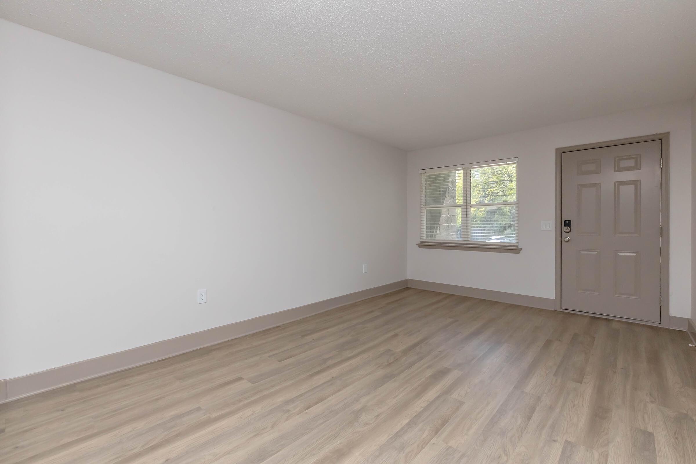 An empty room with light-colored walls and wooden flooring. There is a window on one side allowing natural light in, and a beige door with a handle on the adjacent wall. The space appears clean and minimalistic, suitable for various uses.