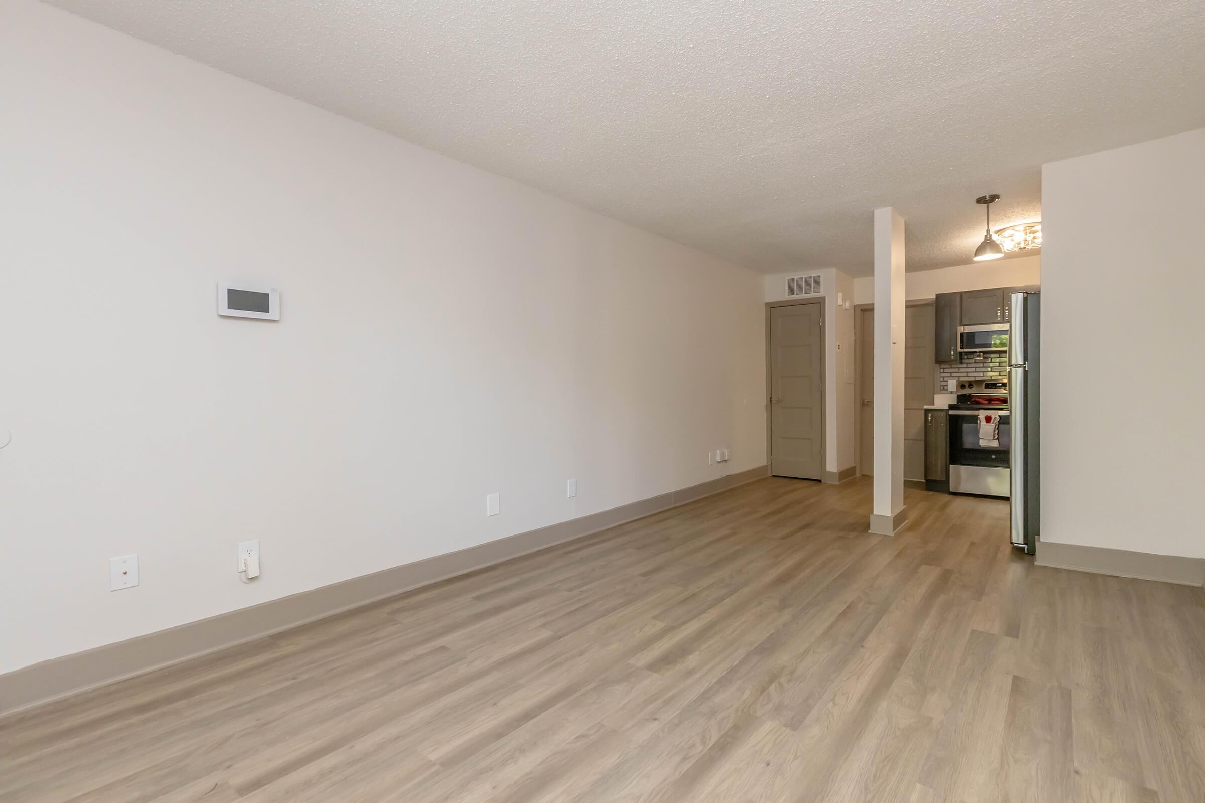 Spacious living area in an apartment with light-colored walls and modern laminate flooring. A wall-mounted thermostat is visible on the left. In the background, a doorway leads to a kitchen featuring gray cabinets and stainless steel appliances, illuminated by a chandelier above.