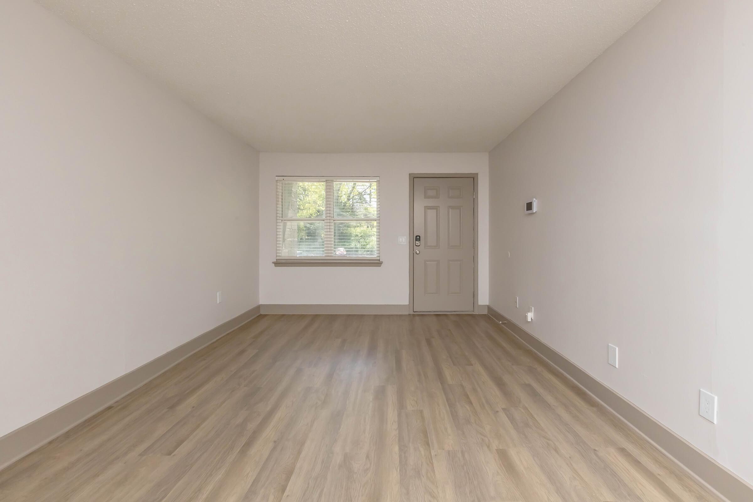 A light-filled, empty room with a beige wall color and light brown laminate flooring. There is a single window on the left, offering a view outside, and a closed door on the right leading to another area. The room has minimal decor and feels spacious, suitable for various purposes.