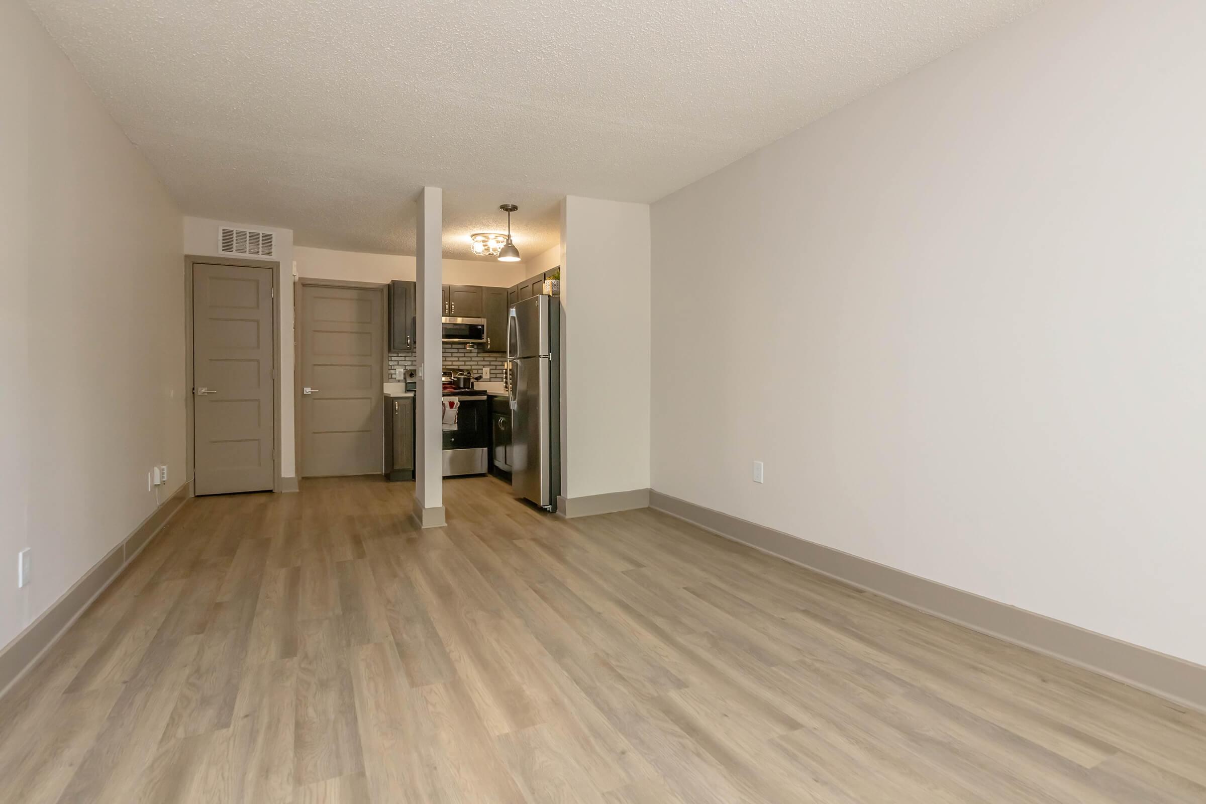 Interior view of a modern apartment showcasing a spacious living area with light wood flooring. There are gray walls, a doorway leading to another room, and a kitchen visible in the background with stainless steel appliances. The space is well-lit and features a minimalist design.