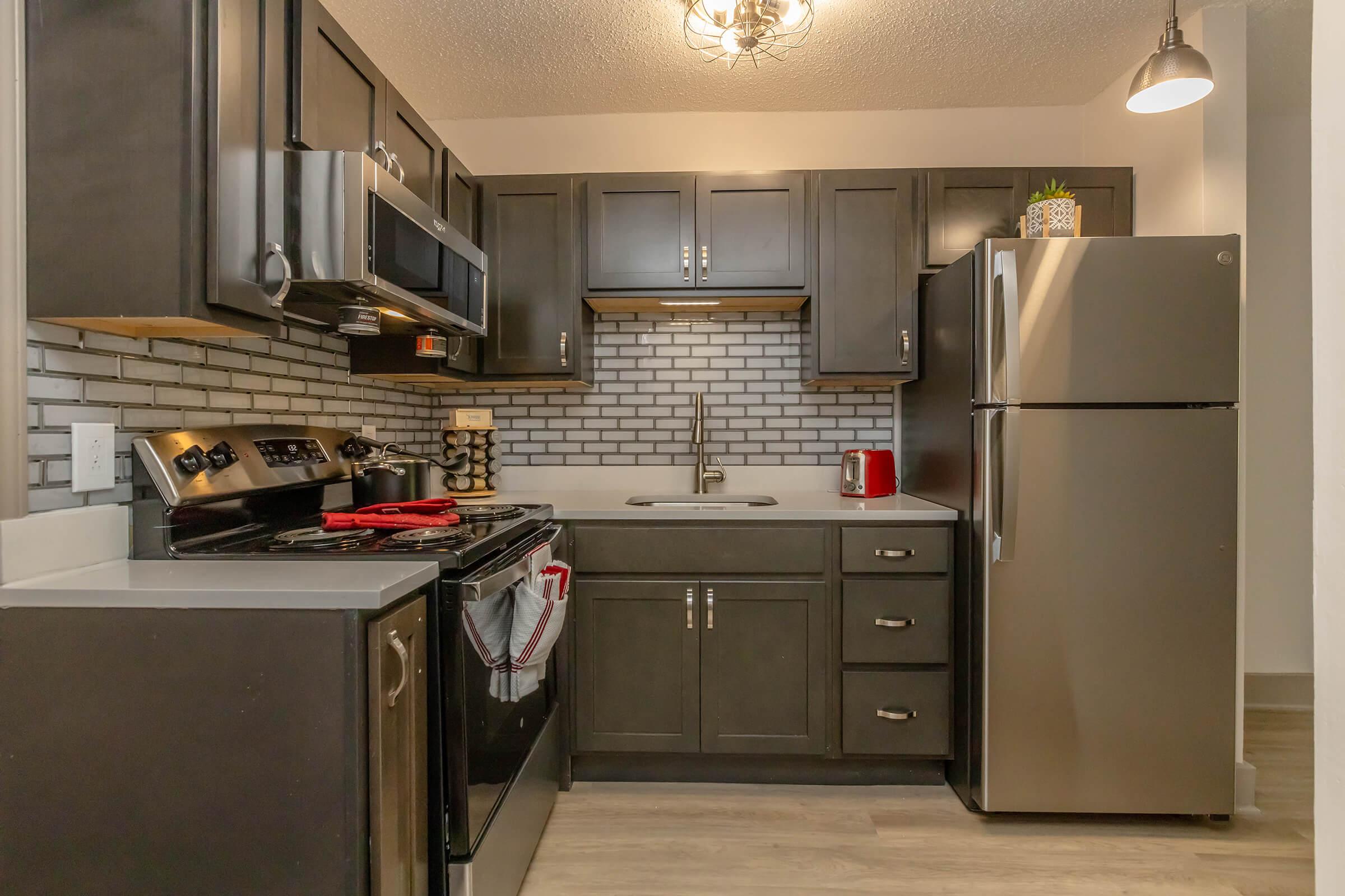 Modern kitchen featuring dark wood cabinetry, stainless steel appliances, and a gray tile backsplash. The layout includes a stove, refrigerator, and sink, with red accents and a small potted plant on the countertop. Dim lighting adds a cozy atmosphere.