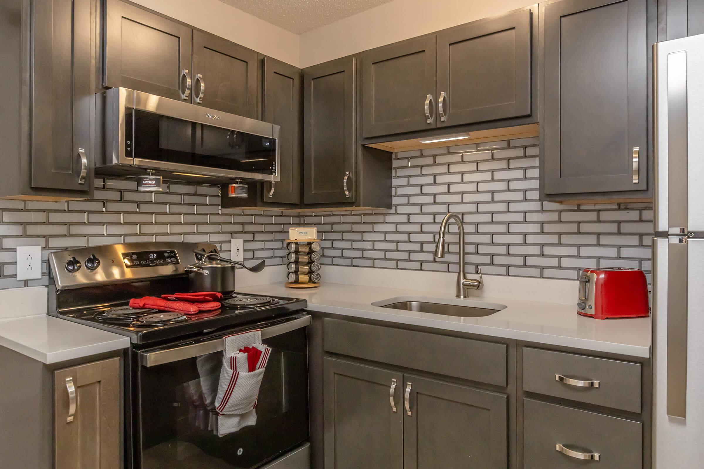 Modern kitchen featuring dark cabinets, a stainless steel microwave, and stove. A subway tile backsplash complements the sleek design. Countertop holds a sink and kitchen appliances, including a red toaster and spice rack. Bright lighting enhances the contemporary style, creating an inviting cooking space.
