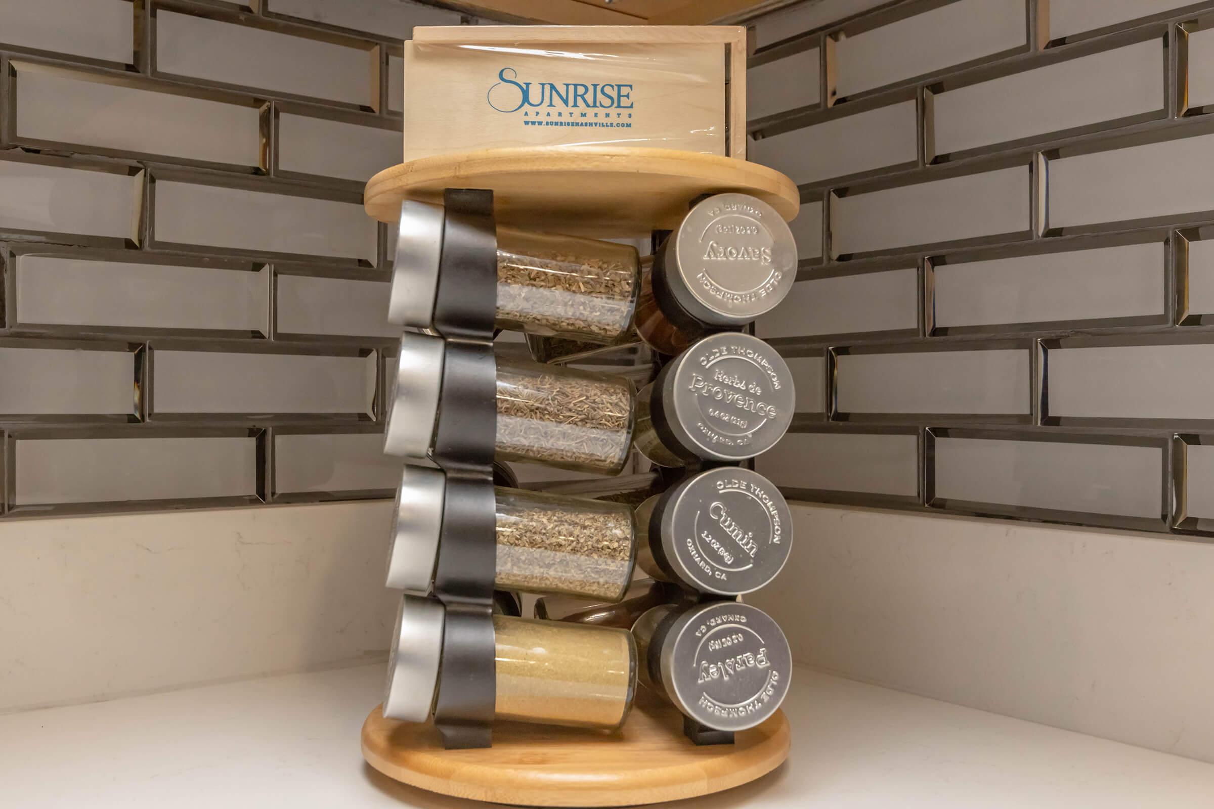 A wooden spice rack holding six glass jars filled with various spices. The rack is circular and displayed on a kitchen countertop with a tiled backsplash. The top of the rack features a brand logo that reads "Sunrise."