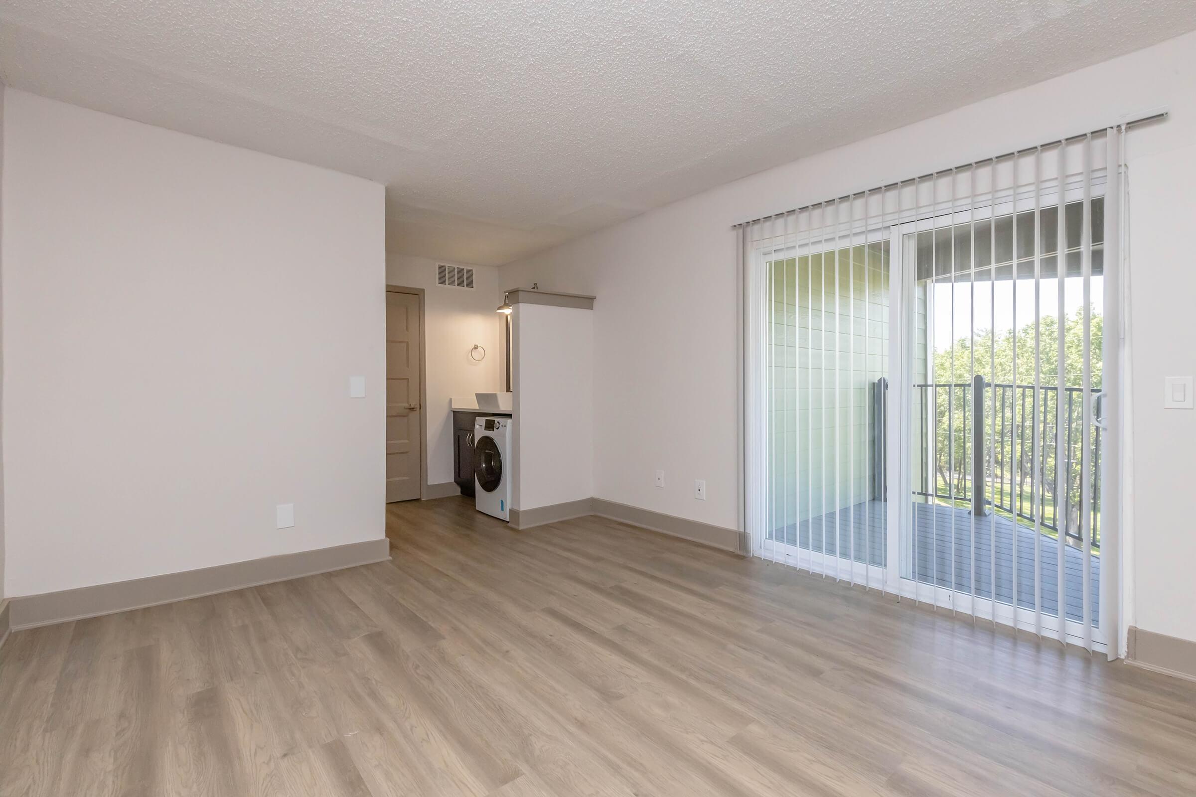 Spacious room with light-colored walls, laminate flooring, and a sliding glass door leading to a balcony. A washer and dryer are located in the corner near a doorway, with natural light streaming in from the balcony, creating a bright and airy atmosphere.