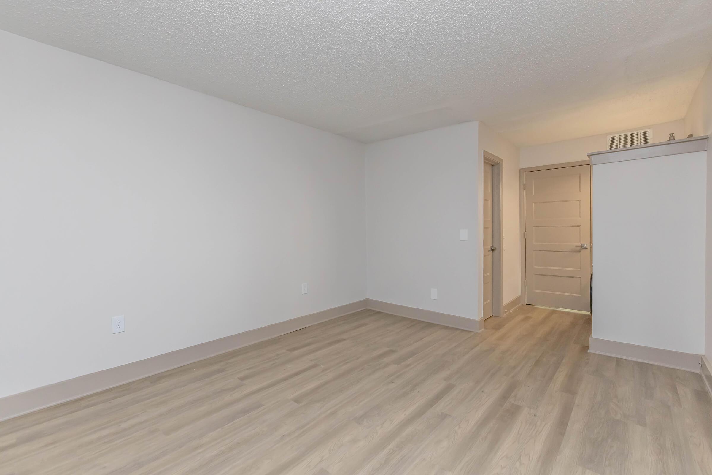 A spacious, empty room with light-colored walls and a textured ceiling. The floor is covered in light wood-like laminate. A doorway is visible on the right, leading to another space, and there are no furnishings present, creating a clean and minimalist appearance.
