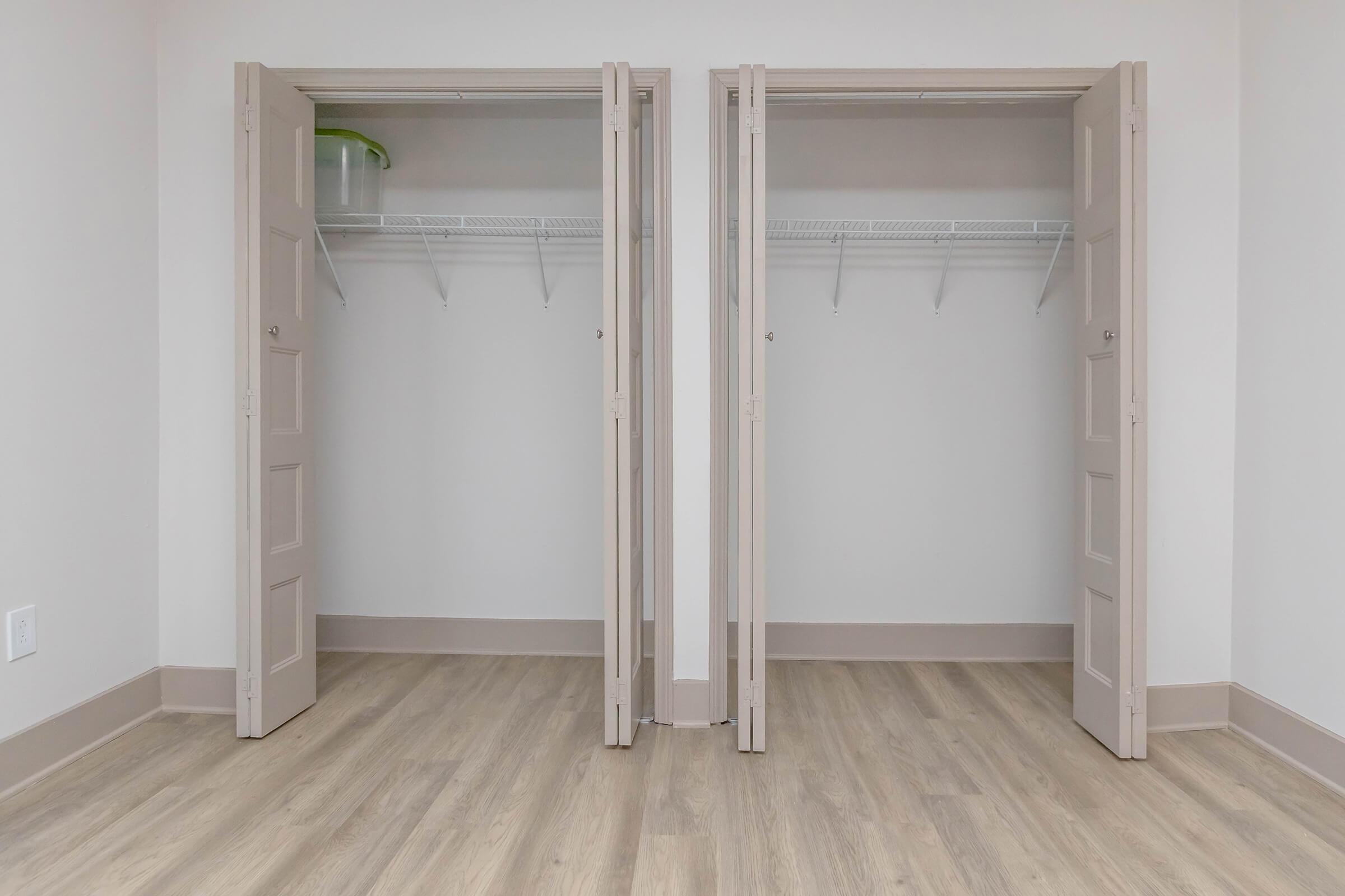 Two empty closets with double doors, featuring wire shelving inside. The walls are painted light gray, and the floor is covered with light wood laminate. The setting appears to be a well-lit, clean room with neutral colors.
