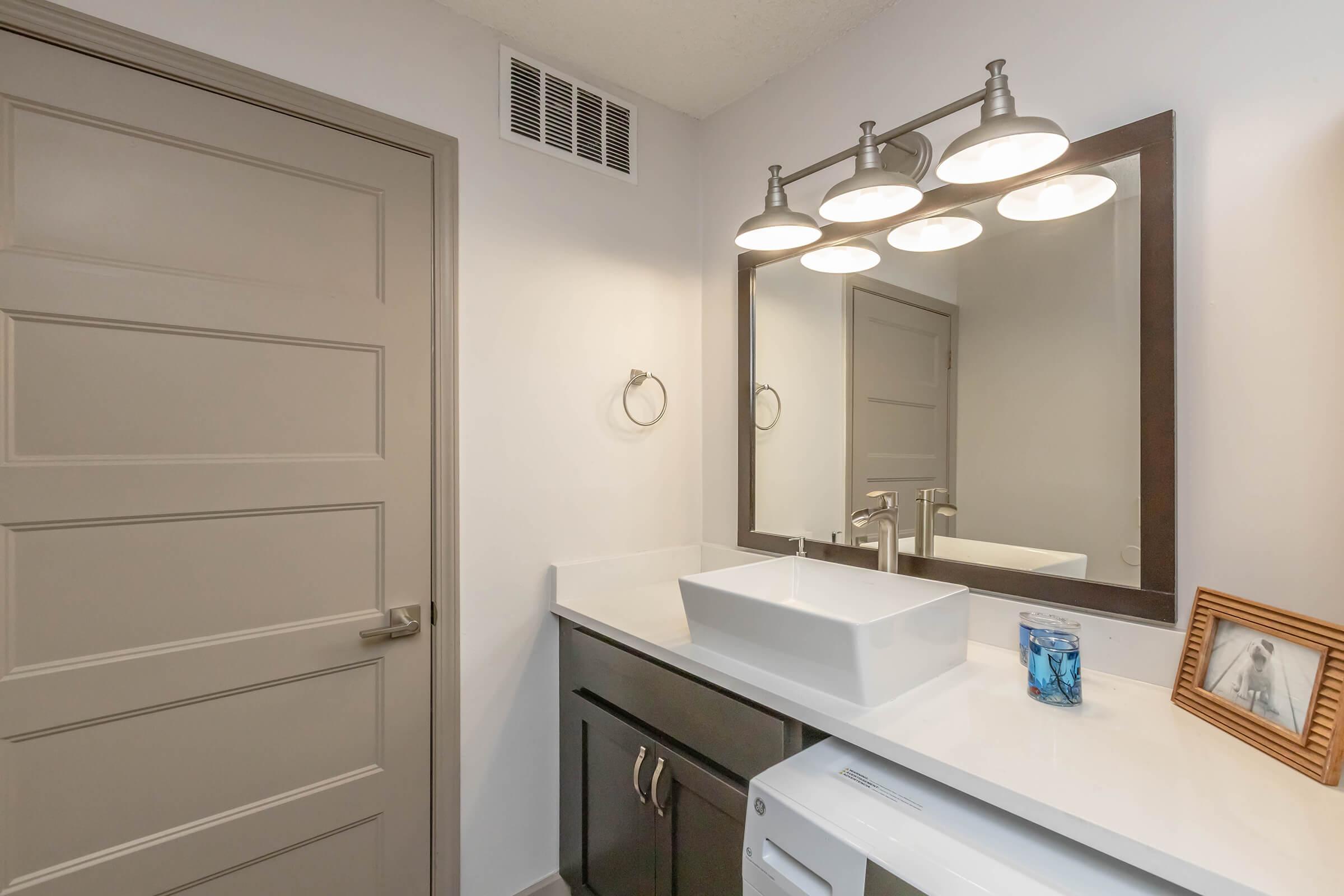 A modern bathroom featuring a sleek white countertop with a square sink, a large mirror with four light fixtures above, dark cabinetry, and a closed door. On the counter, there is a decorative blue glass item and a framed photo. The walls are painted in a light color, creating a fresh and clean atmosphere.