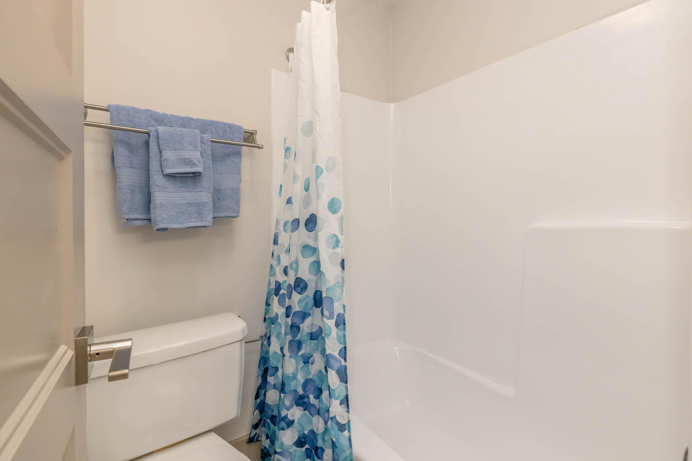 A small bathroom featuring a white bathtub and a shower with a colorful blue and white polka dot curtain. There are neatly folded blue towels hanging on a silver towel bar next to the shower, and a white toilet is positioned beside the tub. The walls are painted a light neutral color.