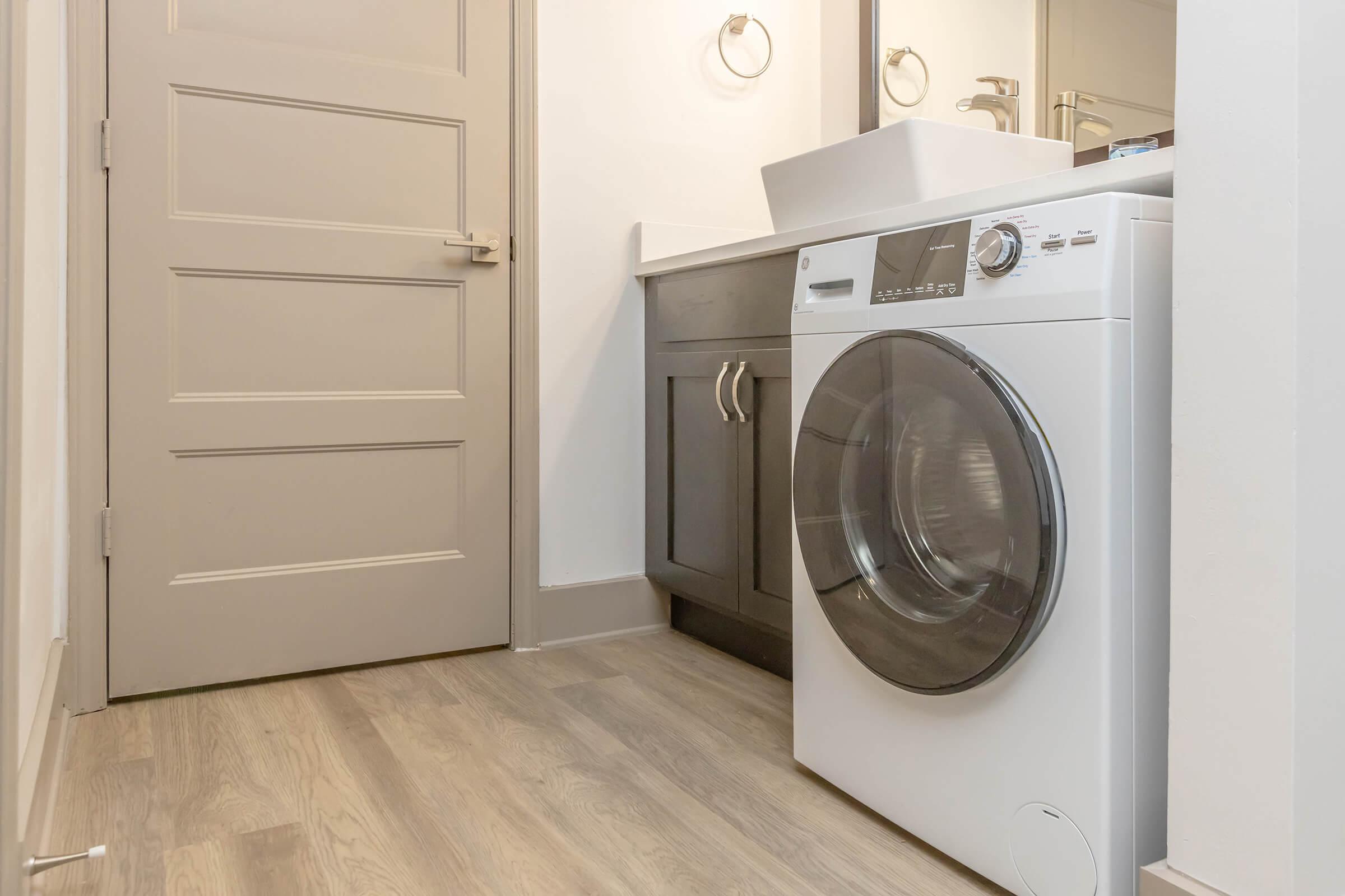 A modern laundry room featuring a white washing machine with a transparent door, positioned next to a sleek gray cabinet and a minimalist sink. The walls are light-colored, and there’s a wooden floor, creating a clean and contemporary aesthetic. A door is visible in the background.