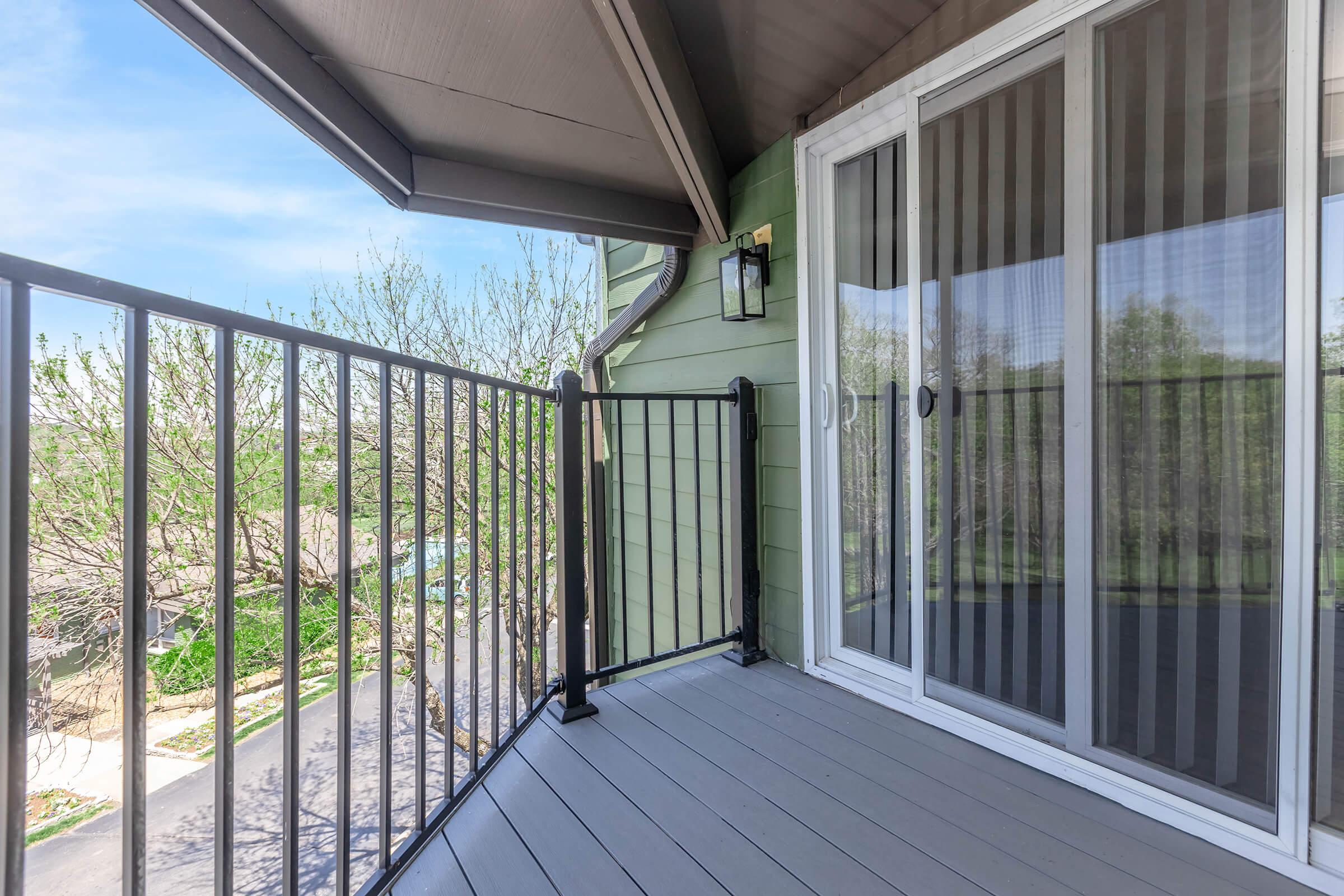 A view of a balcony featuring a gray wooden floor, white railings, and sliding glass doors with sheer curtains. It overlooks a green landscape with trees in the background and a clear blue sky, creating a serene outdoor space.