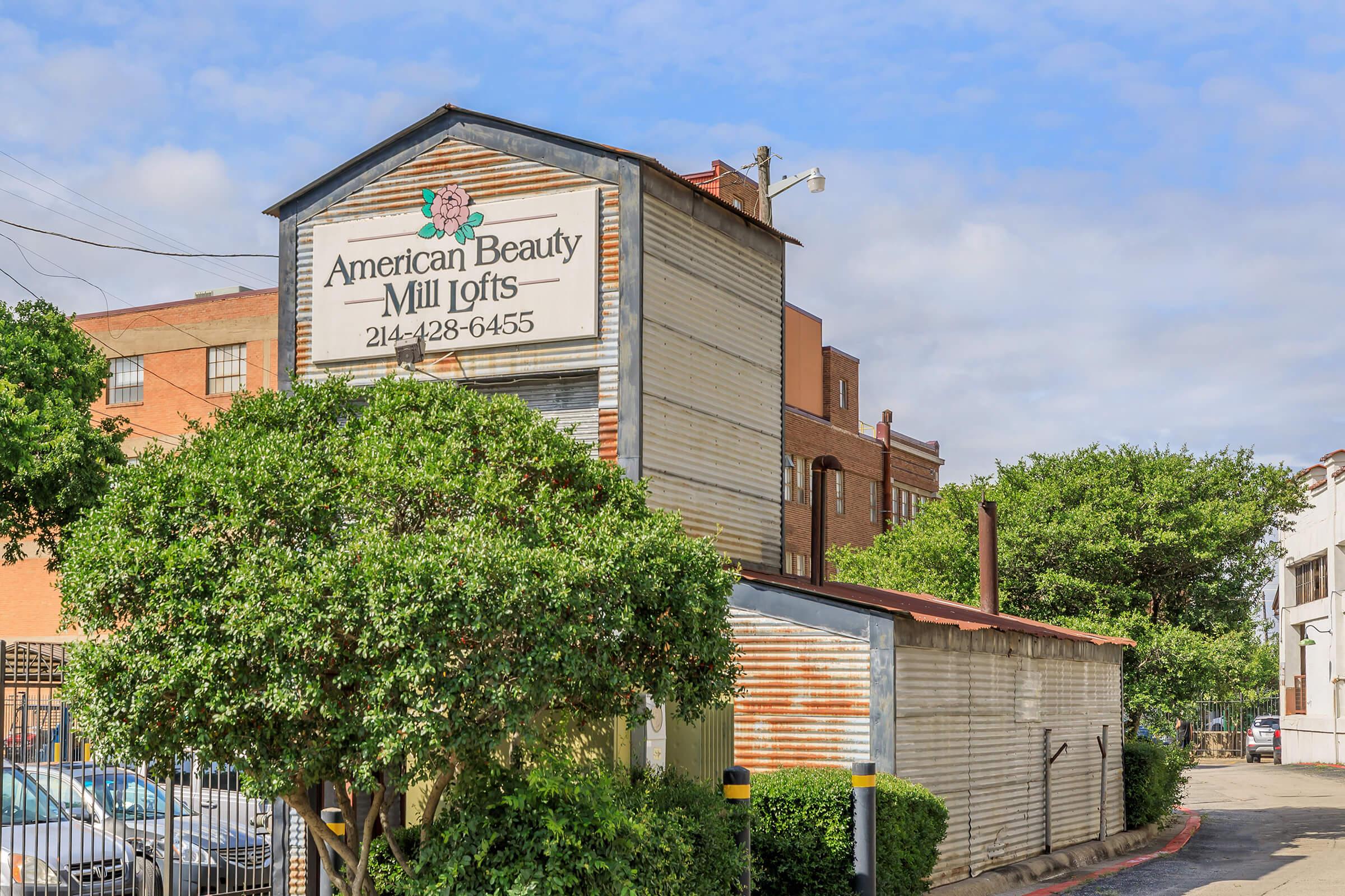 A rustic building with a sign reading "American Beauty Mill Lofts" prominently displayed. The structure has a weathered appearance, with a mix of metal and wooden siding. Lush green shrubs surround the base, and the sky above is partly cloudy. A fence is visible in the background, along with other buildings.