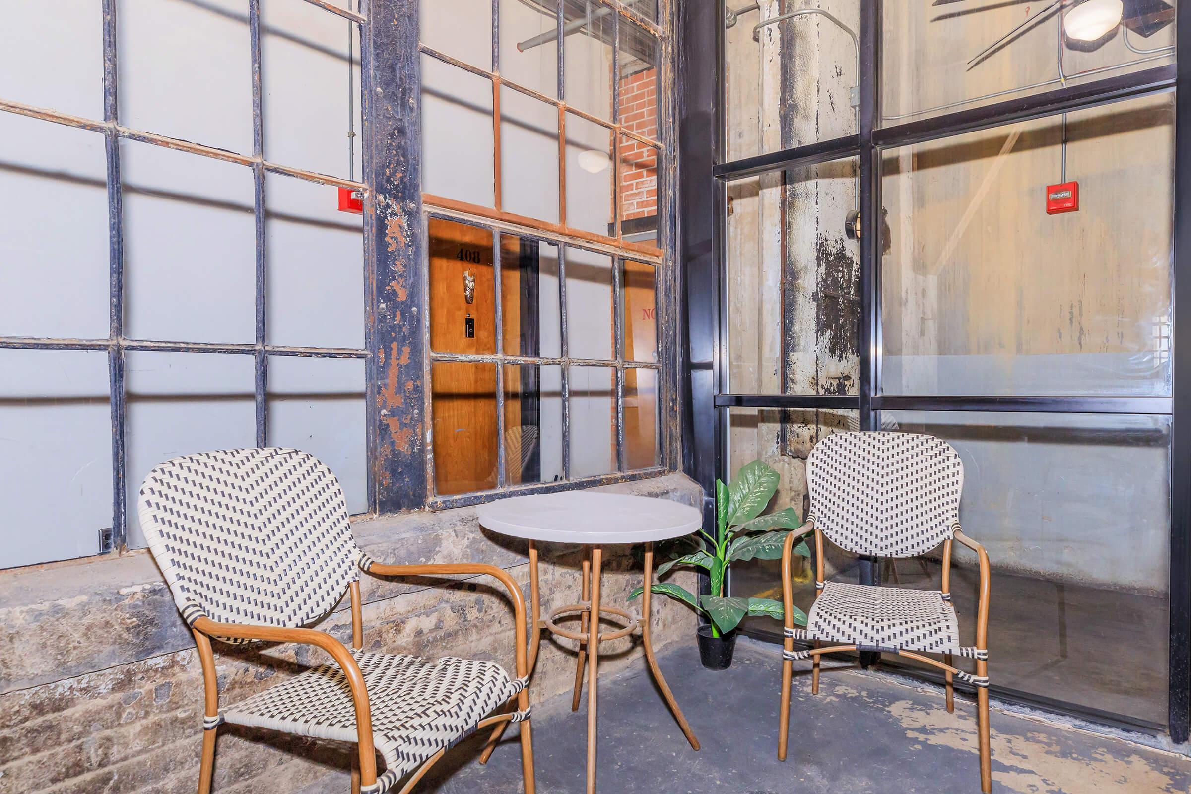 A cozy seating area featuring two patterned chairs and a small round table, set against an industrial backdrop of large windows and exposed brick walls. A small potted plant adds a touch of greenery to the space.