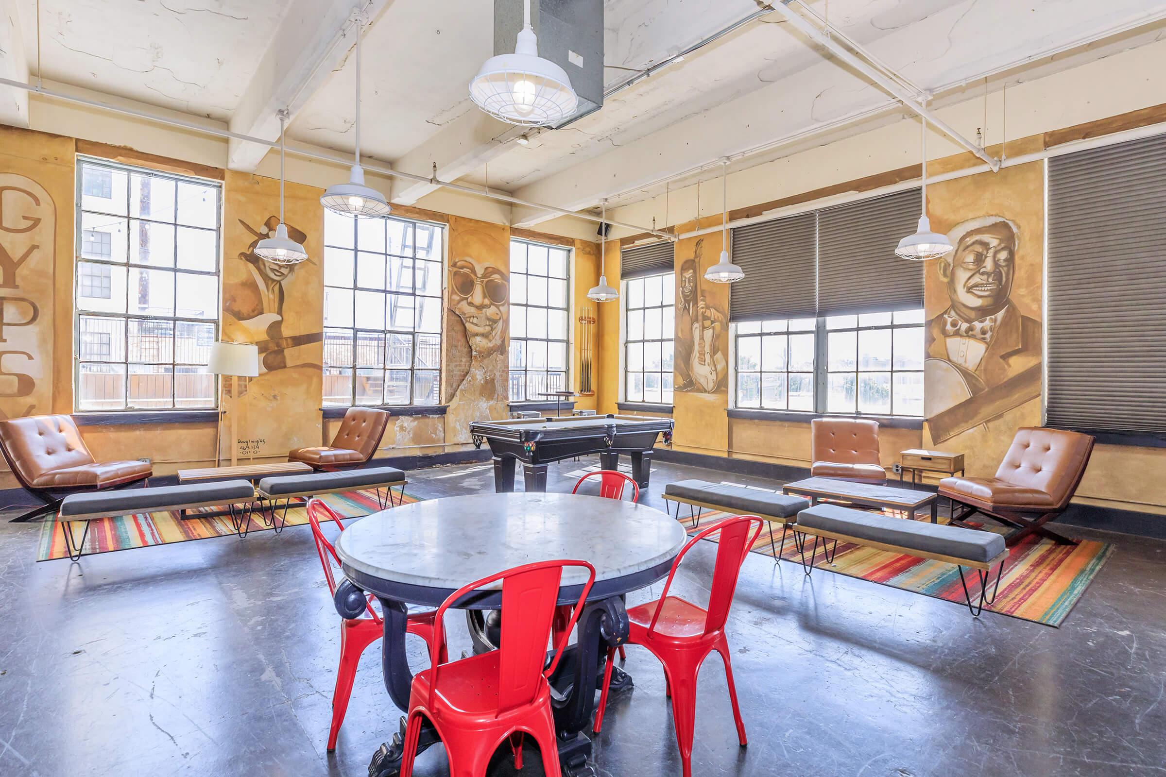 Modern lounge area featuring a round table with red chairs, surrounded by benches. The room has large windows allowing natural light, and the walls are adorned with murals. Two pendant lights hang from the ceiling, and there's a pool table in the background. The floor is dark, and a colorful rug adds warmth to the space.