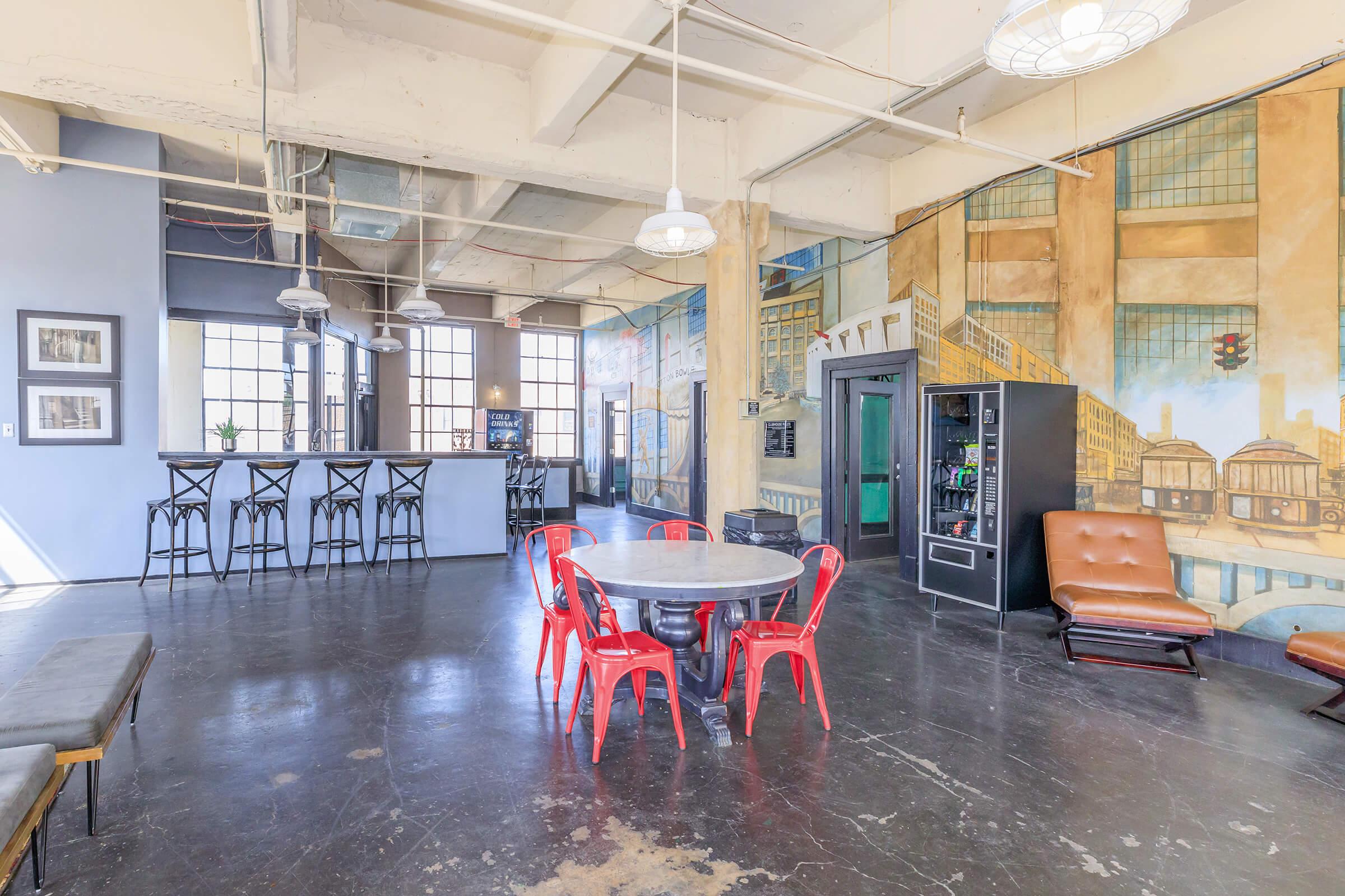 A spacious interior view of a modern loft-style room featuring a round table with red chairs, a brown couch, and bar stools. The walls are decorated with large murals depicting an urban scene. Large windows allow natural light to fill the space, enhancing the open and inviting atmosphere.