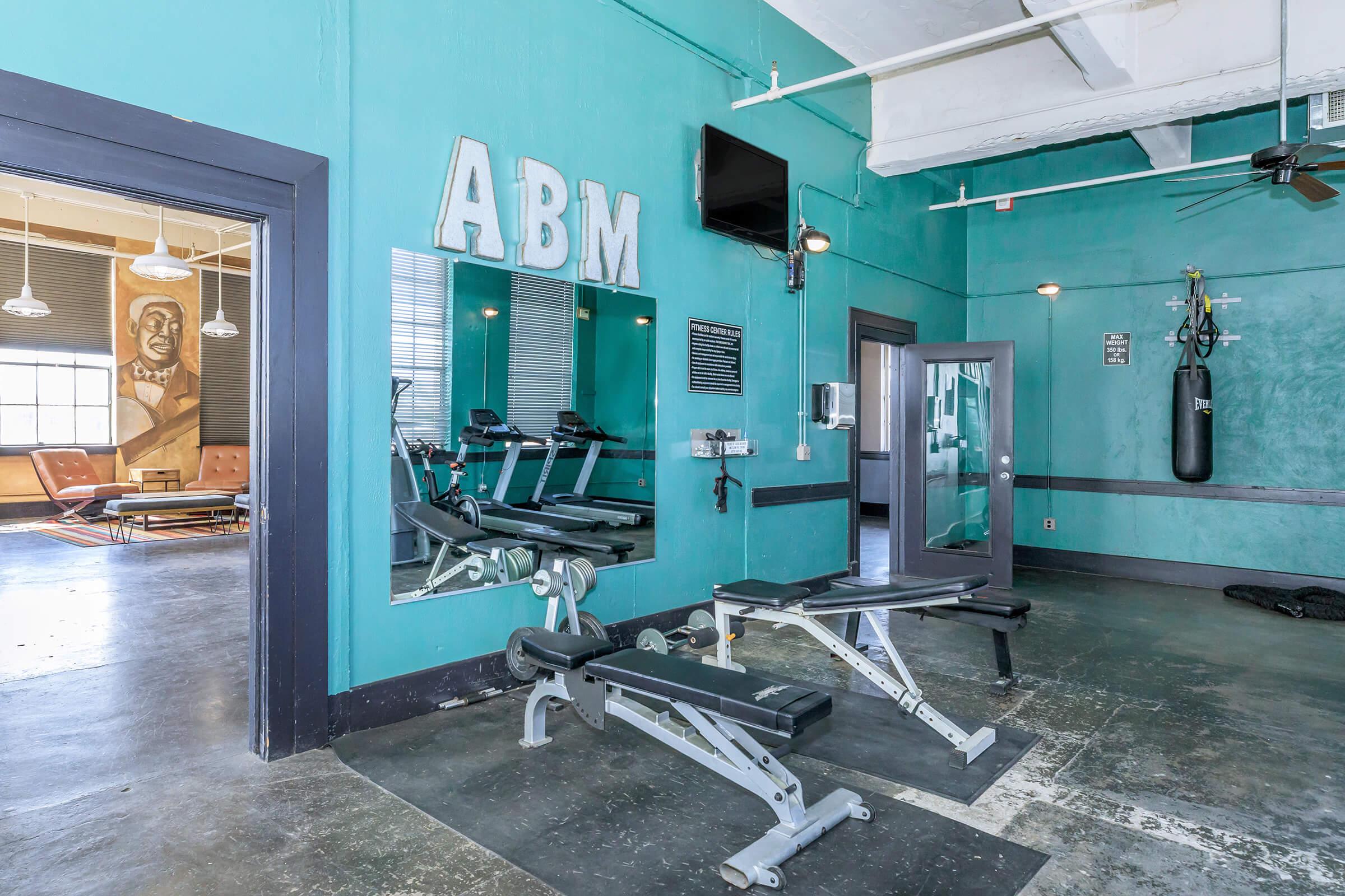 Interior of a gym with teal walls, featuring exercise equipment like benches and mirrors. A television is mounted on the wall, and there’s a punching bag hanging nearby. The space is well-lit, showcasing a modern and clean workout environment.