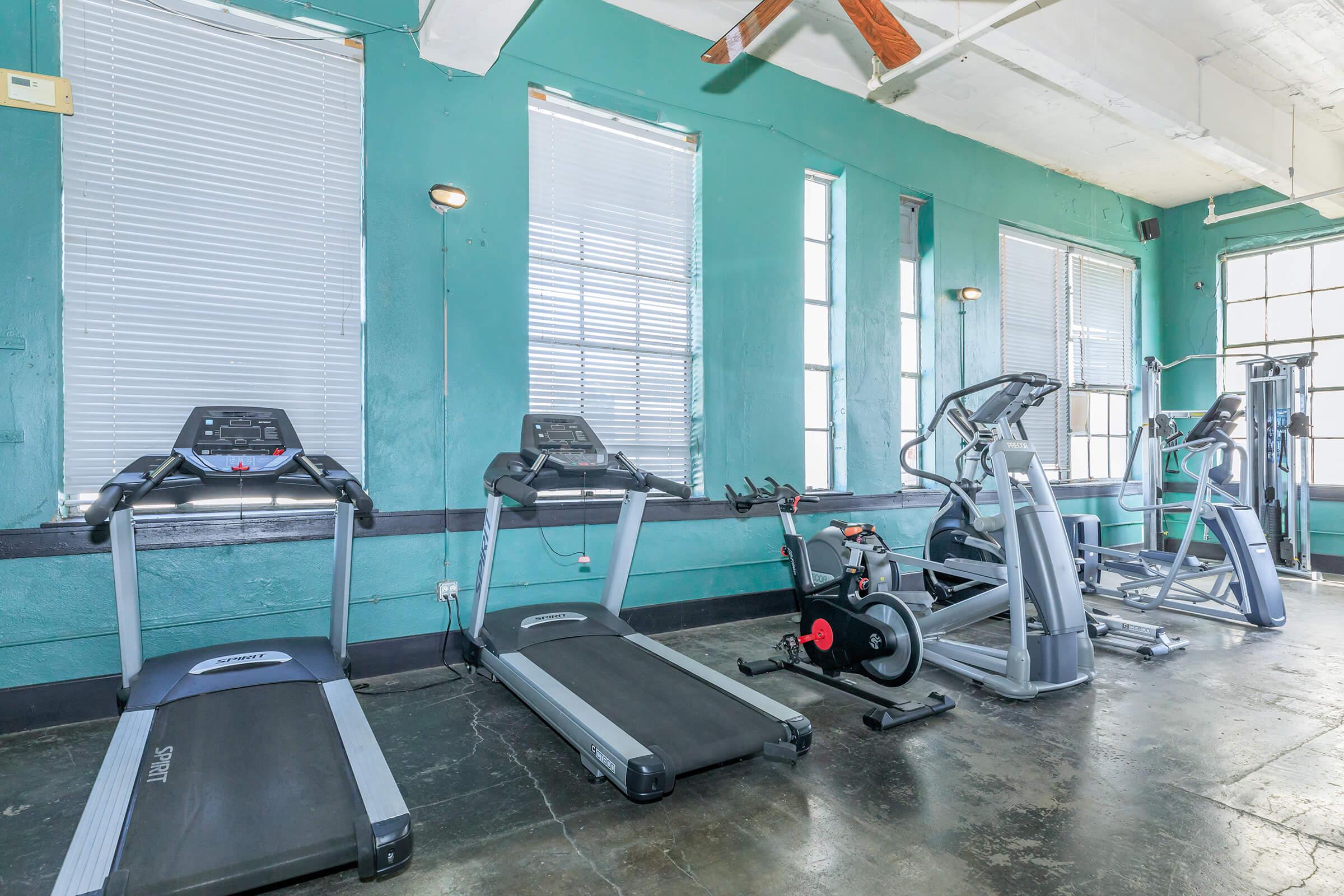 A fitness room featuring several exercise machines including treadmills, an elliptical trainer, and a stationary bike. Large windows provide natural light, and the walls are painted in a teal color. The flooring is dark and polished, creating a modern workout environment.
