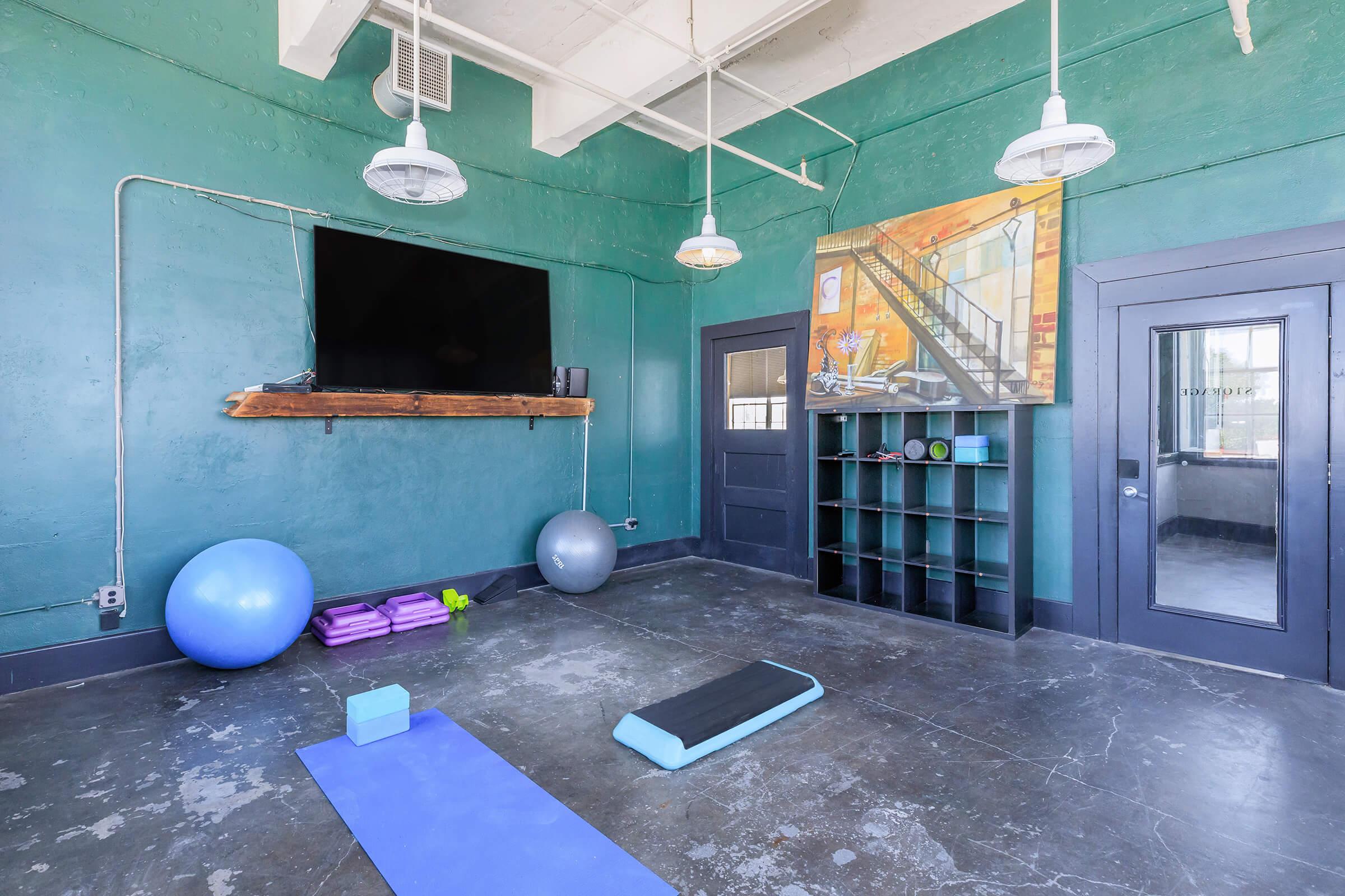 A spacious, modern fitness room featuring green walls, a large wall-mounted TV, and overhead pendant lights. The room contains exercise mats, yoga blocks, stability balls, and a shelf organizing foam weights. A door leads to another area, enhancing the room's functionality for workouts or classes.