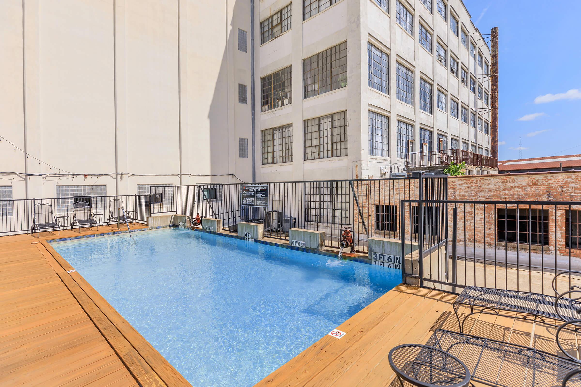 A serene swimming pool surrounded by wooden decking, located in an industrial-style outdoor area. Tall buildings with large windows are visible in the background, and there are metal chairs and tables nearby, creating a relaxing atmosphere. Clear blue skies add to the tranquil setting.
