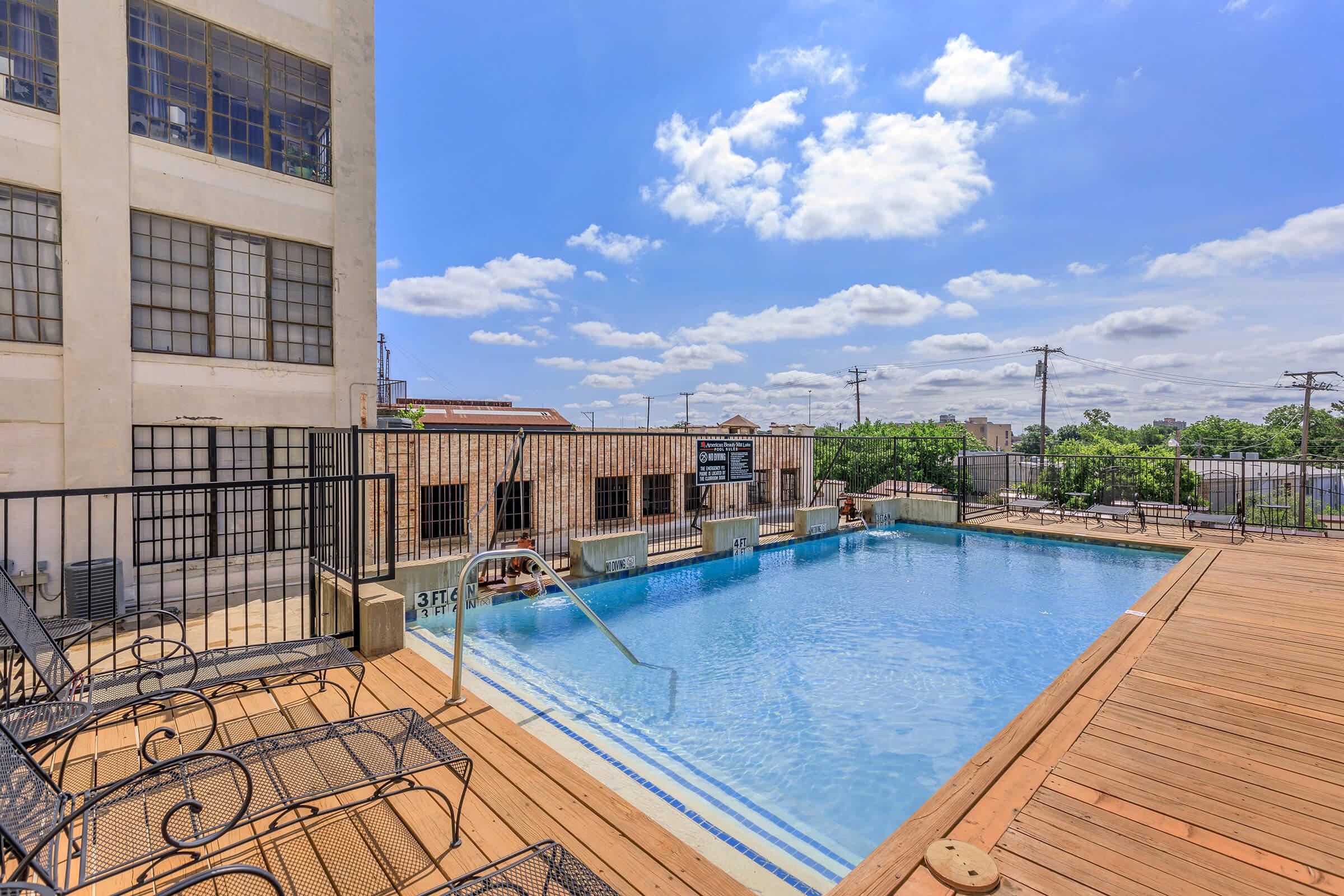 A sunny outdoor swimming pool surrounded by a wooden deck, with lounge chairs and a staircase leading into the water. The backdrop features a large brick building with multiple windows and a cloudy blue sky above. Power lines are visible in the distance, contributing to an urban atmosphere.