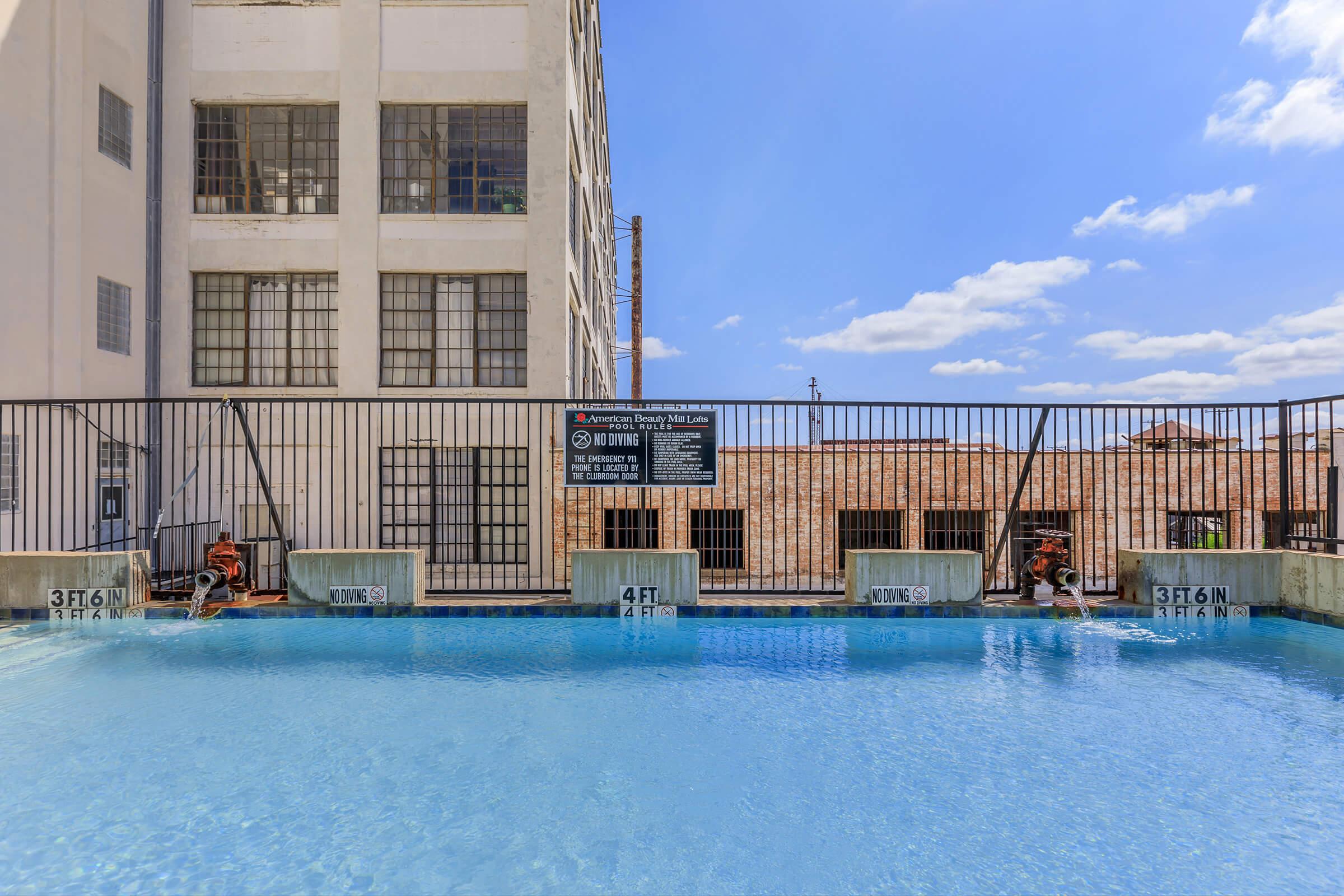Swimming pool with clear blue water and a safety sign, surrounded by a metal fence. The backdrop features a brick building with large windows and a bright blue sky dotted with clouds. The pool has depth markings showing 3 ft and 4 ft increments.