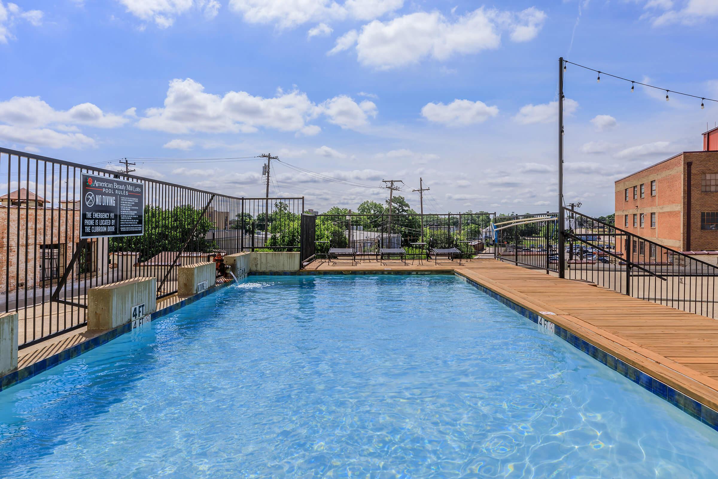 A clear blue swimming pool surrounded by wooden decking. The pool area is enclosed with a black railing, and there is a sign nearby. In the background, there are trees and buildings under a partly cloudy sky, with power lines visible.