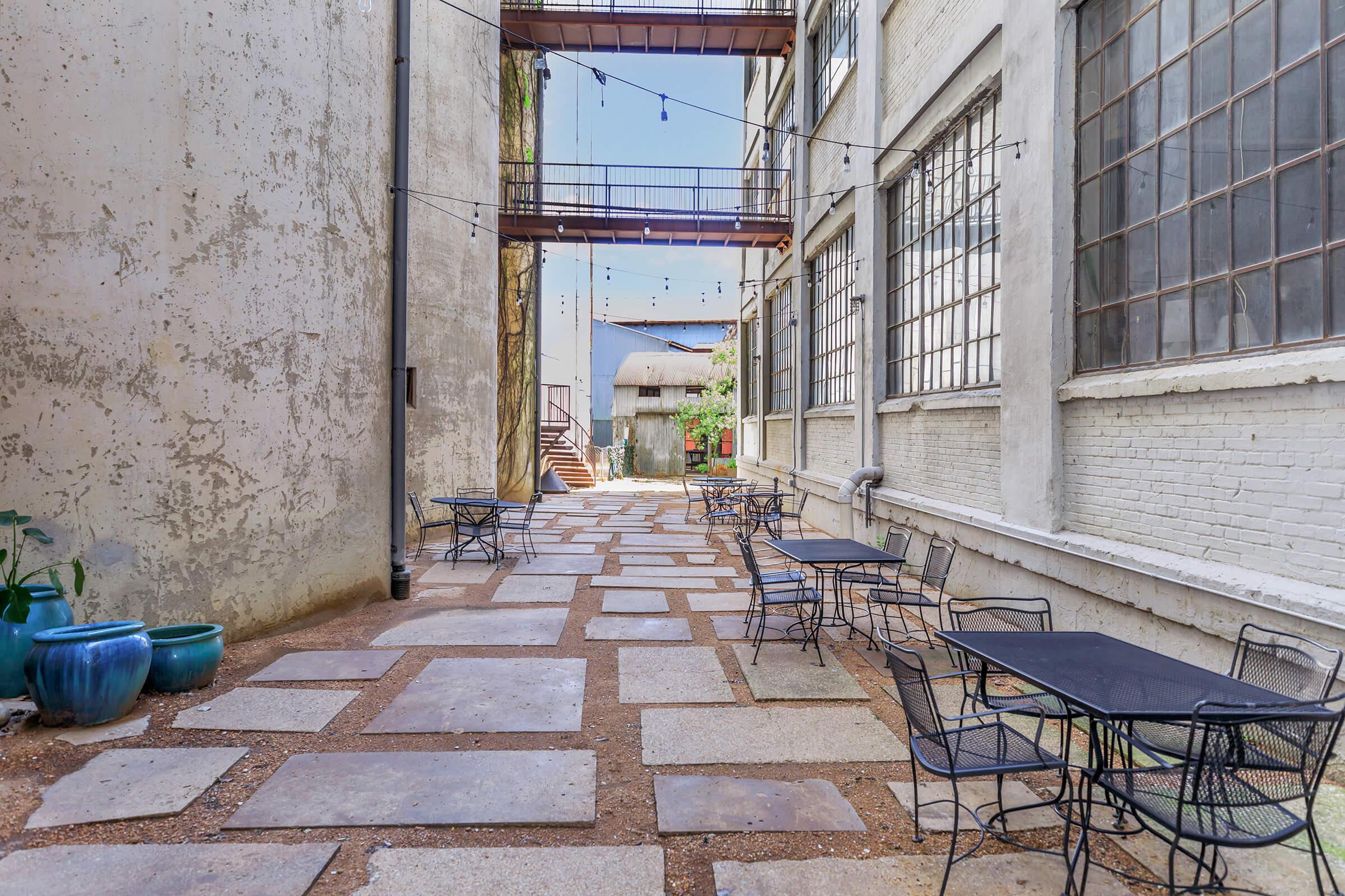 A narrow, sunlit alleyway with a textured stone path, flanked by white, industrial-style walls and large windows. Black outdoor tables and chairs are arranged along the sides, and string lights hang overhead, creating a cozy atmosphere. In the background, a staircase leads to another level.