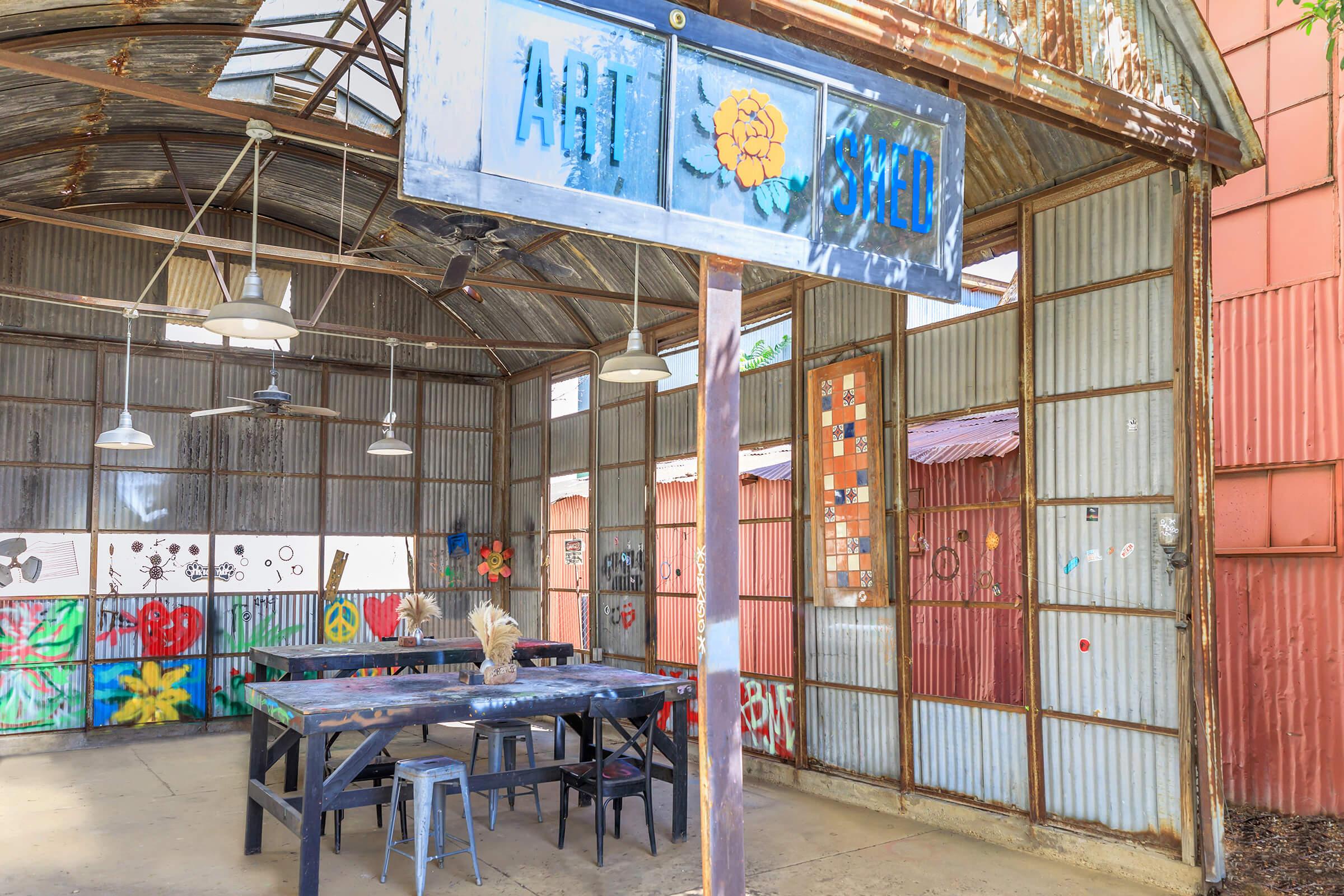 Interior of an art studio with a metal roof and corrugated walls. A sign reading "ART SWELL" hangs above a rustic wooden table surrounded by chairs. Colorful murals and floral designs decorate the walls, creating a vibrant and creative atmosphere. The space is bright with natural light filtering through the windows.