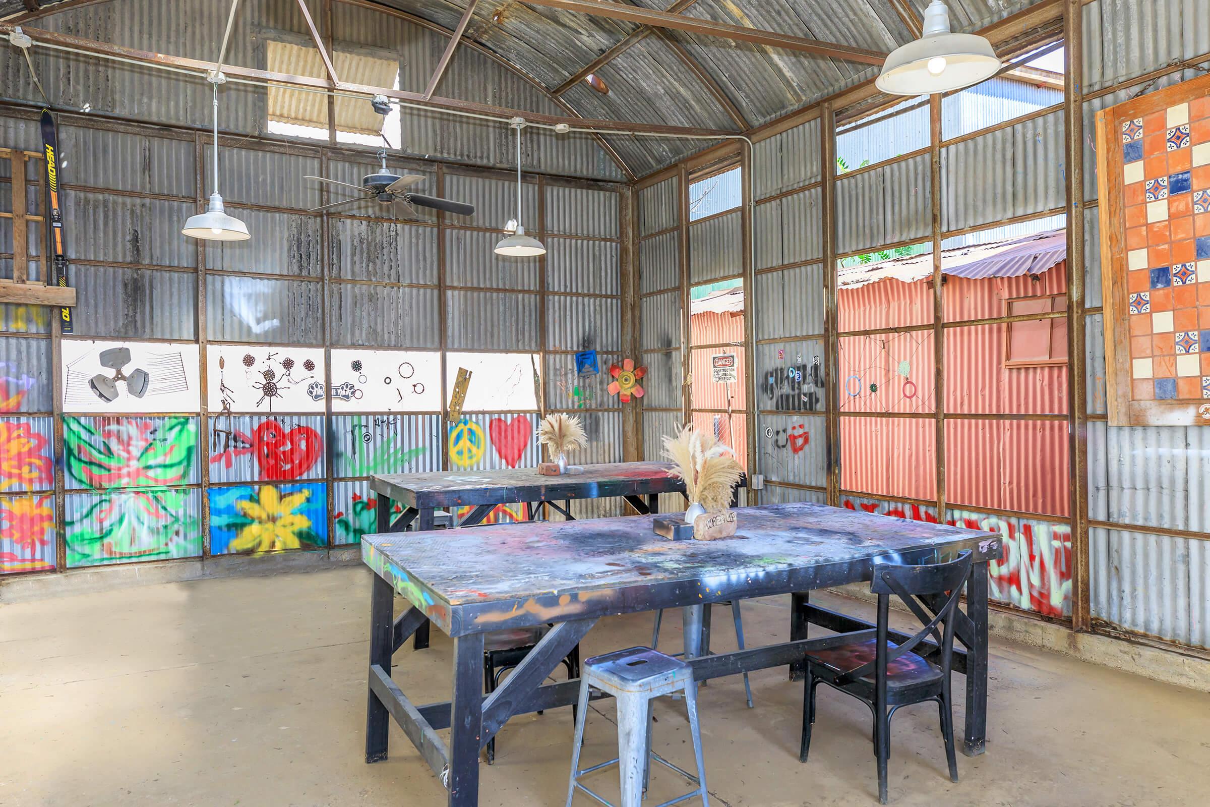 An interior view of an art studio featuring metal walls and a high ceiling. There are two large wooden tables with art supplies, and several artworks displayed on the walls, including colorful flowers and abstract designs. Natural light comes from overhead lamps, creating a vibrant and creative atmosphere.
