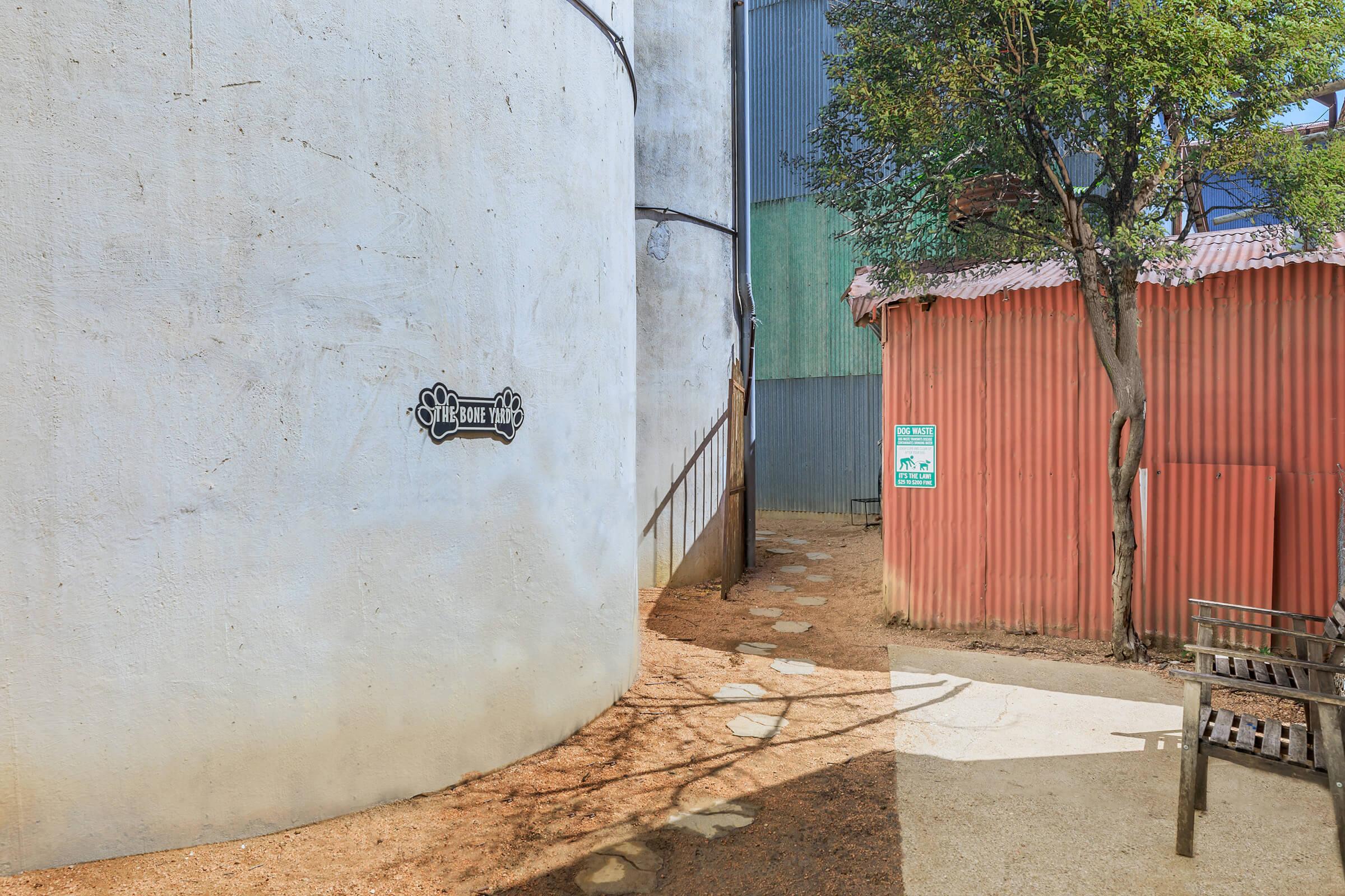 A narrow path leads between two buildings: a round, white structure with a decorative sign and a colorful, corrugated metal building. Sunlight casts shadows on the ground, and a small tree adds greenery to the scene. A wooden bench is visible nearby, along with signs on the walls.