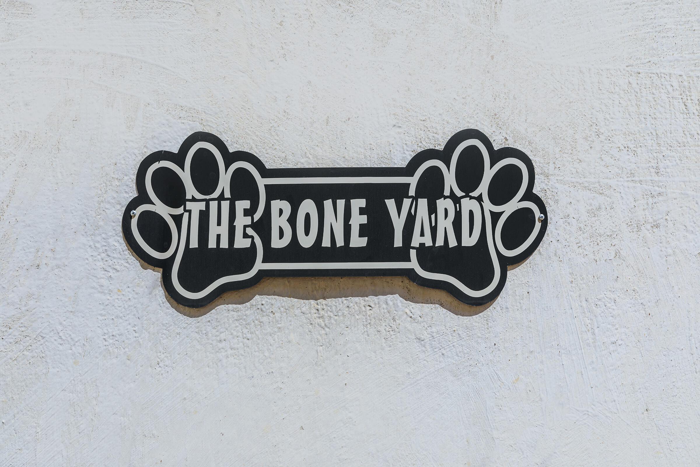 Sign with the words "THE BONE YARD" in bold letters, flanked by two paw print designs, set against a light, textured background. The sign has a playful and welcoming appearance, suggesting a pet-related theme or business.