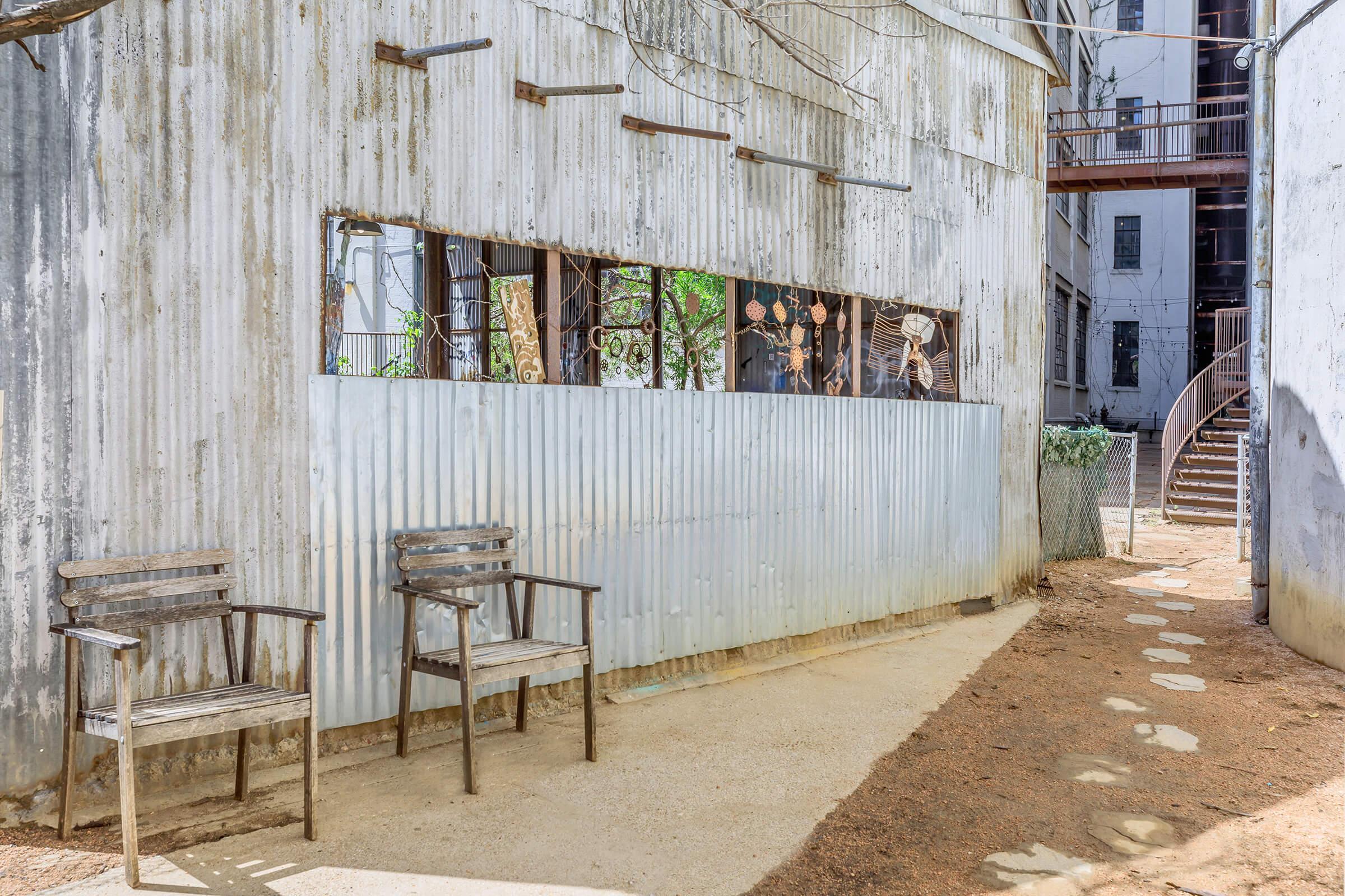 A weathered outdoor area featuring two wooden chairs in front of a corrugated metal wall with large windows displaying artistic cutouts. The ground is sandy with stone path elements leading toward a building in the background.