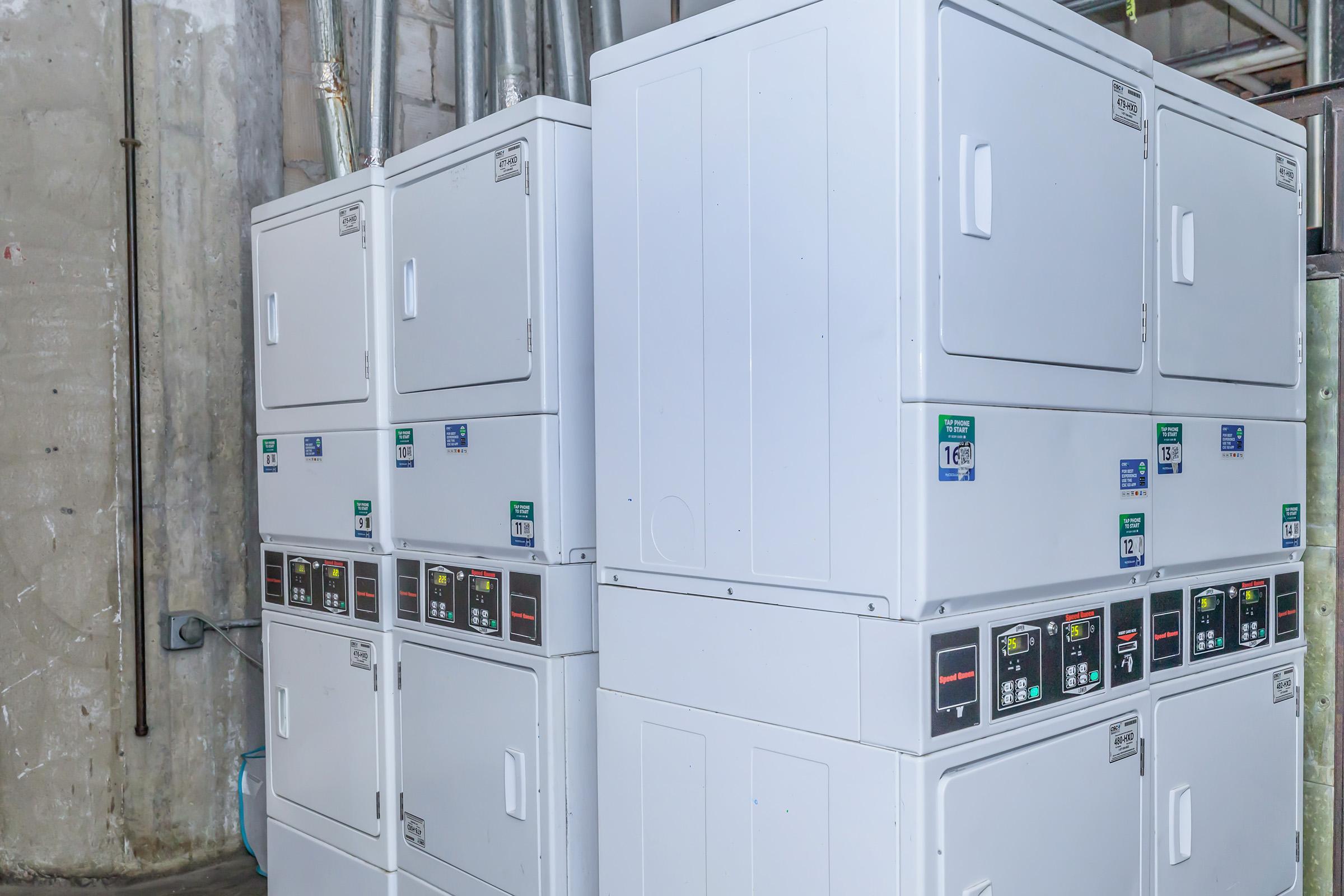 Rows of stacked white commercial laundry machines in a utility area. The machines include both washers and dryers, featuring digital displays and control panels. The background is a gray concrete wall with visible plumbing and electrical installations.
